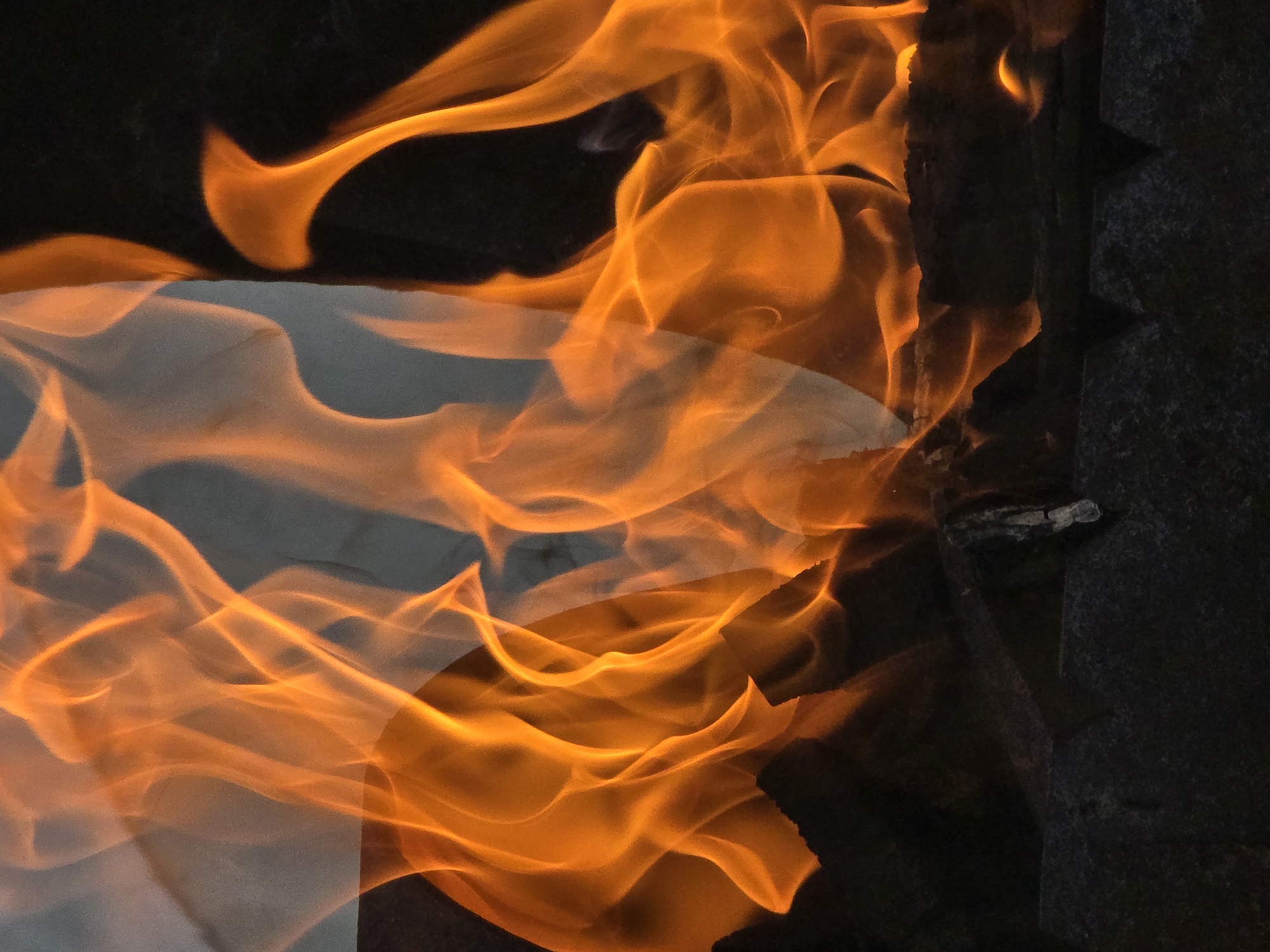 A close-up, eye-level shot shows bright orange flames from a fire. The fire appears to be in a dark, possibly metal, fire pit with some dark charcoal or wood visible. The flames are dynamic, with some parts glowing brighter than others, and they create a sense of movement. A faint, lighter-colored material is visible behind the flames, creating a translucent effect. The background is mostly dark, with the exception of a small reflection of light in the upper right portion of the image.