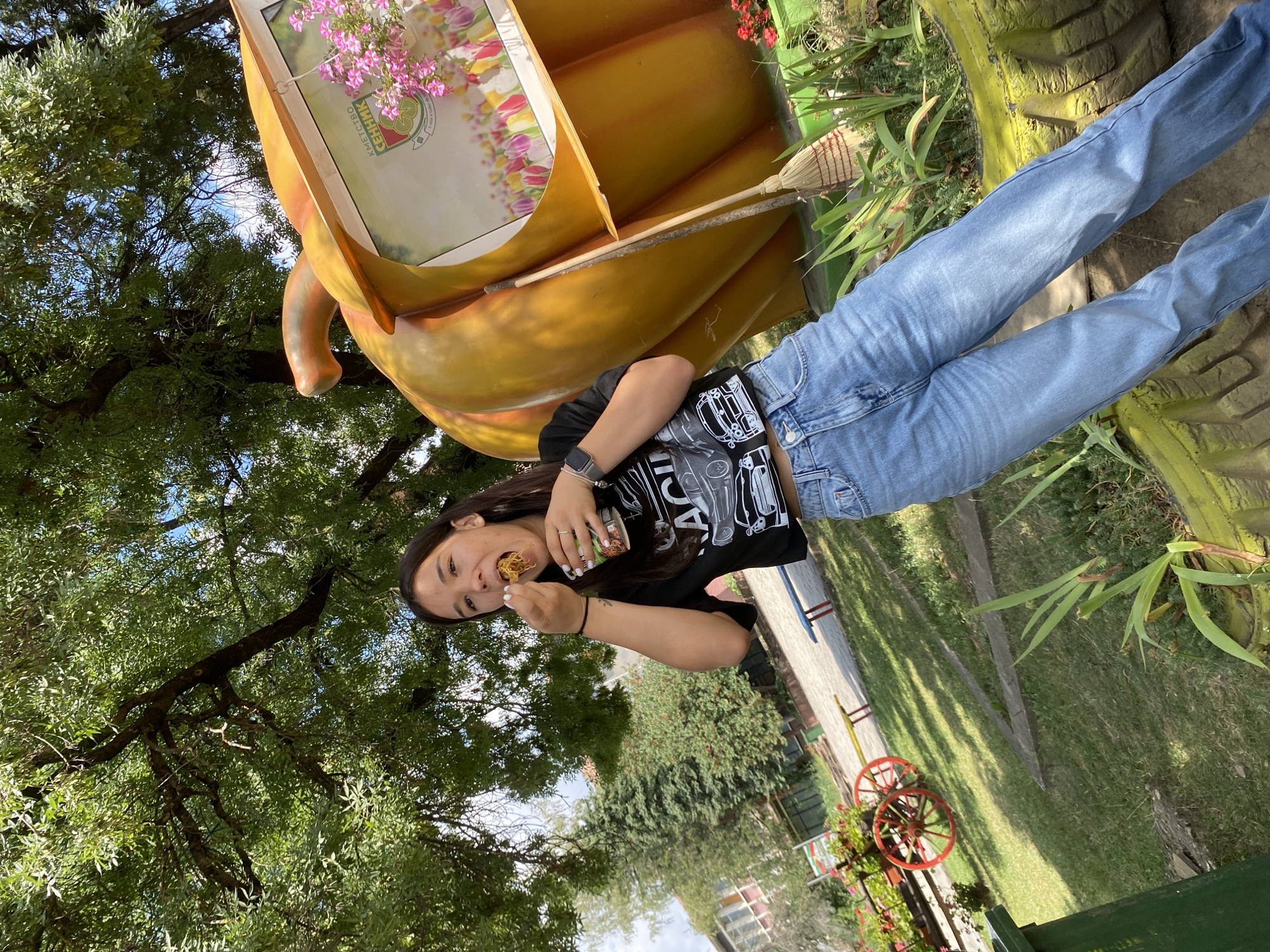 In Sennik, Bulgaria, a young woman with long dark hair, wearing a black graphic t-shirt and light blue jeans, stands outdoors on a sunny day, eating from a cup with a fork. She is positioned in front of a large, orange, pumpkin-shaped structure, which appears to be a decorative installation. The pumpkin has a window-like opening displaying a picture of tulips and a sign with Bulgarian text. To the right of the pumpkin, a straw broom leans against it. The background features lush green trees with dappled sunlight, a grassy area, a decorative wagon with red wheels, and buildings in the distance. The scene is set in a park or a public outdoor space.