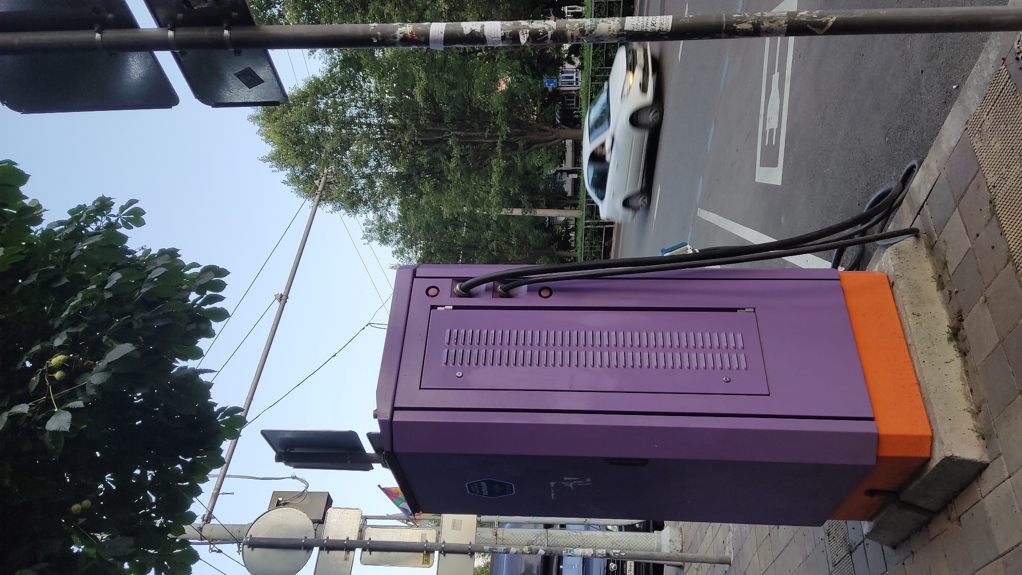 This is a medium shot, eye-level outdoor photograph of a purple electric vehicle charging station situated on a paved sidewalk in Krasnodar, Russia. The charging station, which has an orange base, is positioned next to a street where a white car is driving by, appearing blurred due to motion. Overhead, tree branches with leaves and some green conkers hang above the scene. Traffic lights and poles with signage are visible in the background. The sky is clear and blue. The overall environment suggests an urban setting.