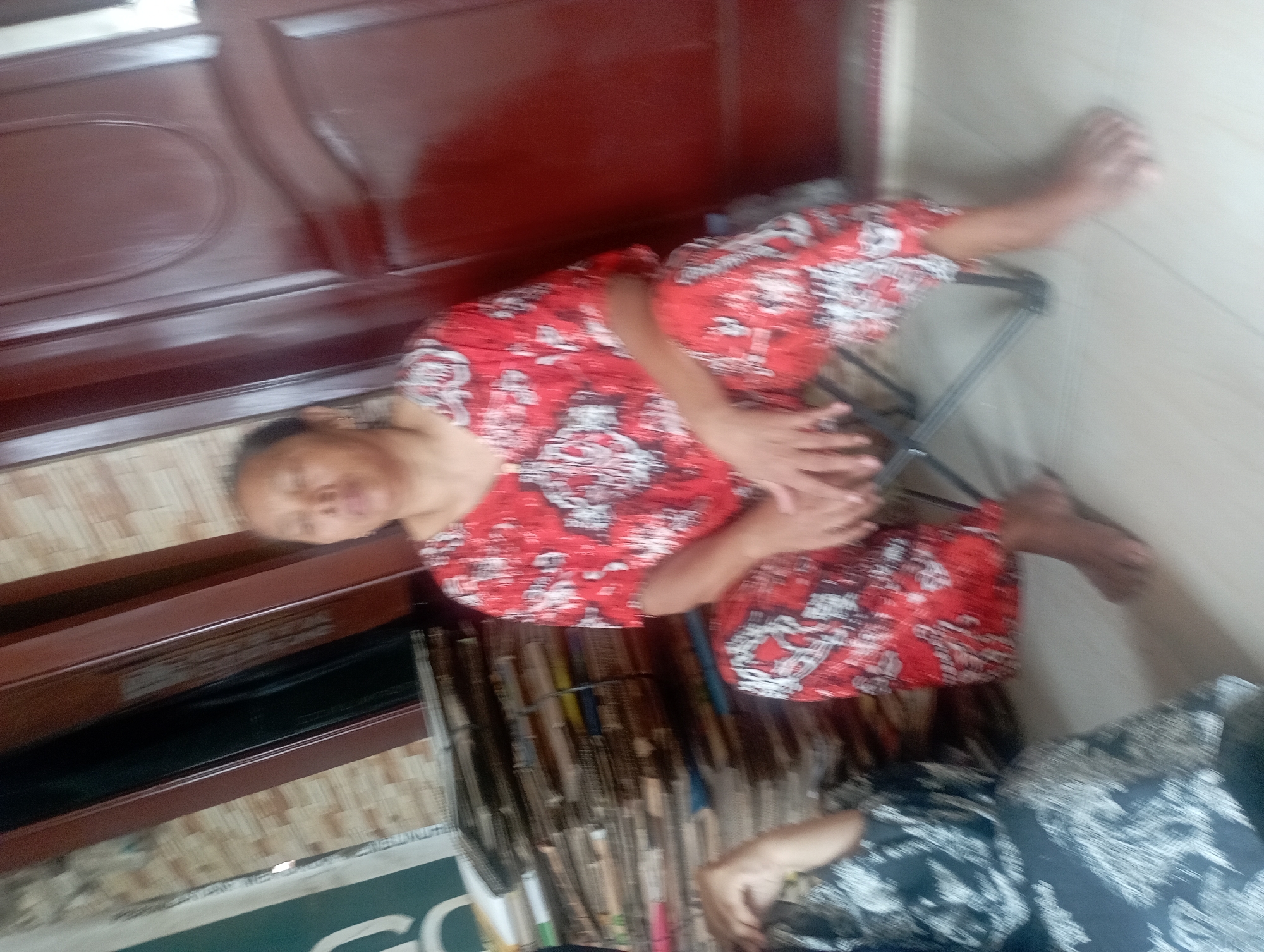 In a room, likely in Maos Kidul, Indonesia, a woman sits on a folding chair wearing a red and white patterned outfit. She has short dark hair and appears to be looking down. To her left, another person, partially visible, is seated and also wearing a dark patterned outfit. Behind the seated woman is a large stack of what appears to be cardboard or paper. The room has a tiled floor and a large, dark-colored door is visible to the right of the frame. The wall behind the woman has a textured pattern.