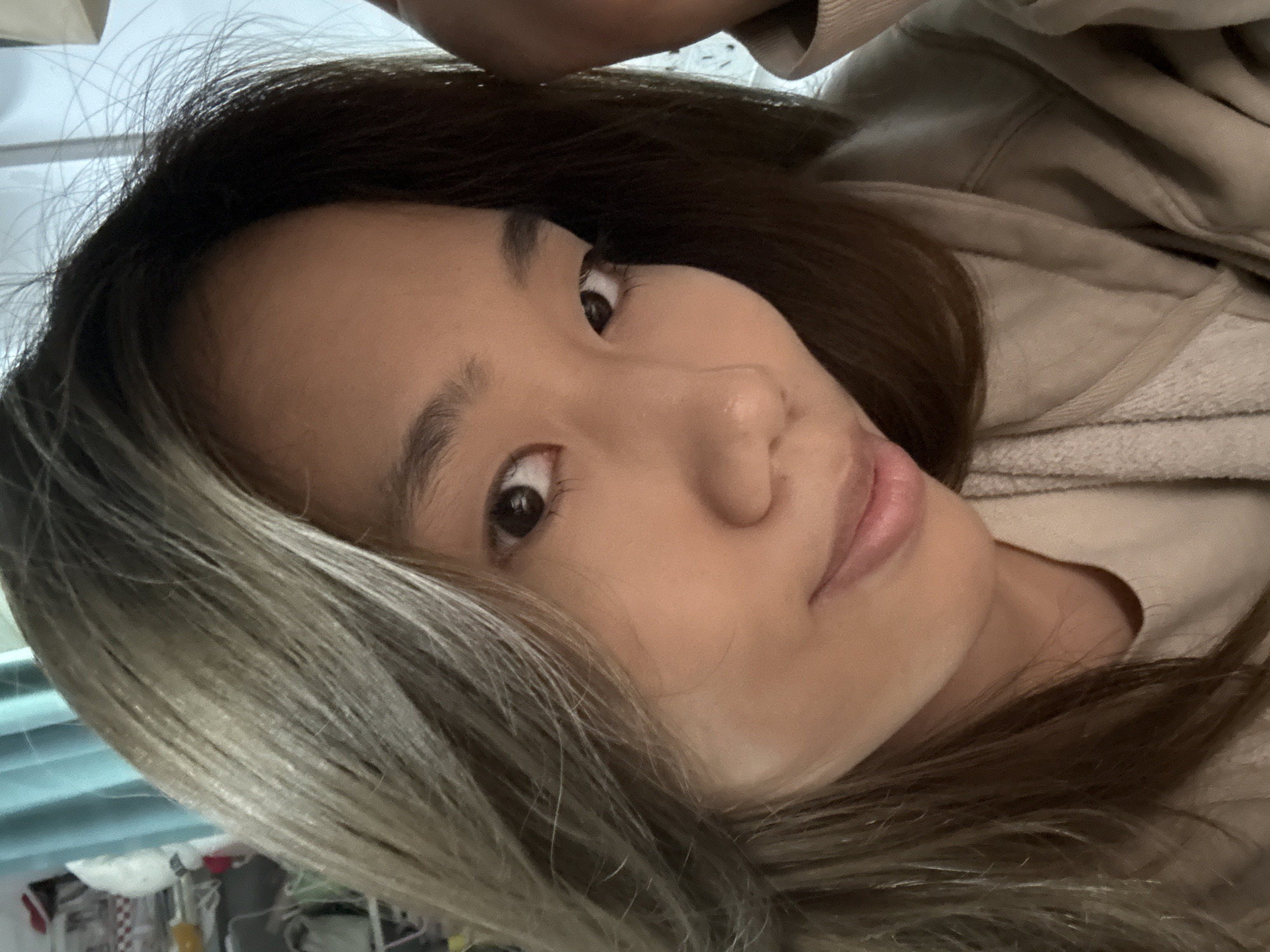 A close-up, slightly angled selfie shows an Asian woman with light brown skin and brown eyes. Her dark brown hair fades to blonde in the lower left corner. She is wearing a beige sweatshirt. The background is blurred, showing a glimpse of a light blue surface and indistinct objects. There's no clear indication this image was taken in Nan'an District, China.