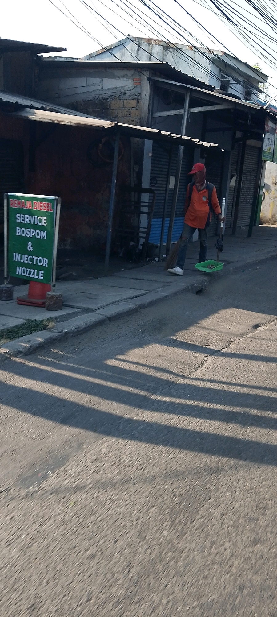 A man wearing a red cap, orange shirt, and blue jeans stands on a sidewalk in front of a shop, holding a broom and a dustpan. He appears to be in the process of sweeping. The shop has a green sign that reads "REMAJA DIESEL SERVICE BOSPOM & INJECTOR NOZZLE". The background shows buildings with corrugated metal roofs and various wires overhead. The scene is set on a street in Depok, Indonesia, with shadows cast across the road by the low-angled sun.