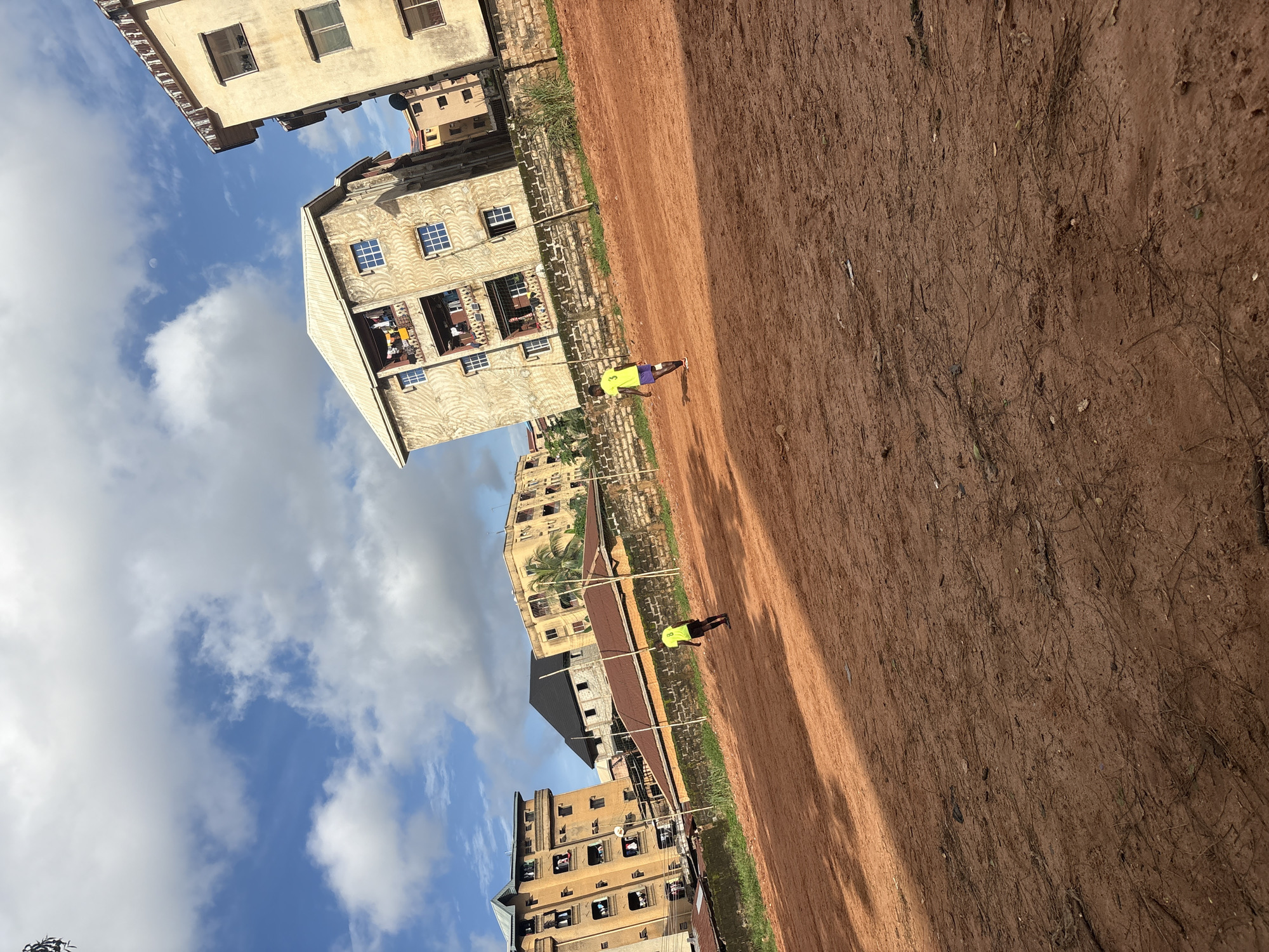 The image depicts an outdoor scene in the city of Onitsha, Nigeria, featuring a reddish-brown dirt field in the foreground. In the background, several multi-story buildings are visible, with the architecture suggesting residential apartments. The sky is partly cloudy, with a mix of blue sky and white clouds, and the sun is casting shadows on the field. Two young males, dressed in athletic attire, are present on the field. One individual, wearing a bright yellow t-shirt and dark shorts, is in the mid-ground, appearing to be in motion. Another individual, similarly dressed in a yellow shirt and purple shorts, is further to the left, also seemingly engaged in an activity on the field. The overall scene suggests a casual outdoor setting, possibly a recreational area within a residential part of the city.