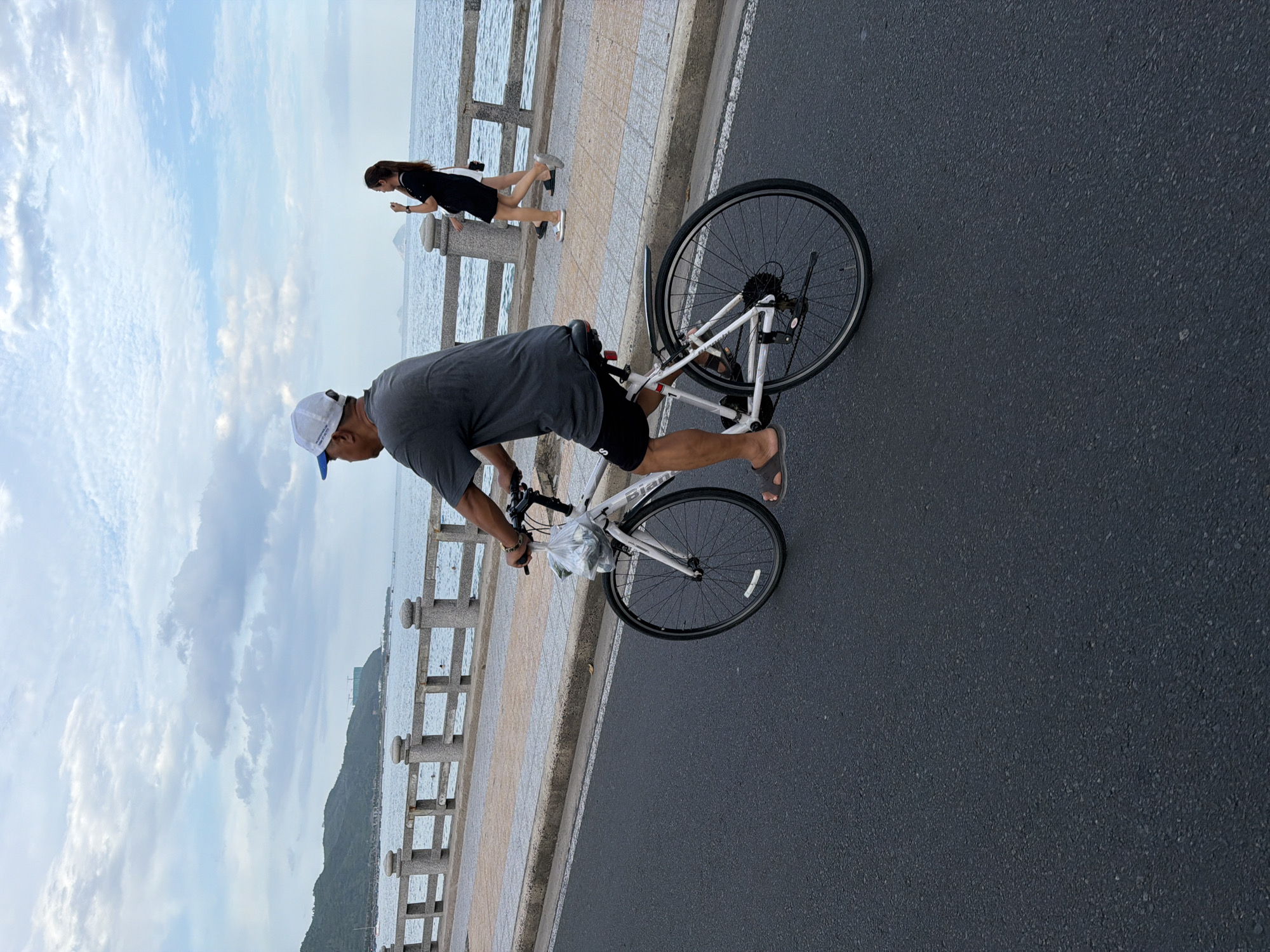 A man is riding a white bicycle on a paved road, his body angled away from the camera. He is wearing a grey t-shirt, black shorts, and a white baseball cap. A plastic bag is hanging from the handlebars of his bicycle. In the background, a woman in a black dress and another person in white are walking on a sidewalk next to a stone railing that borders the sea. The sea is visible beyond the railing, with hills in the distance under a cloudy sky. The scene appears to be along the coast of Nha Trang, Vietnam, likely on a promenade or coastal road.