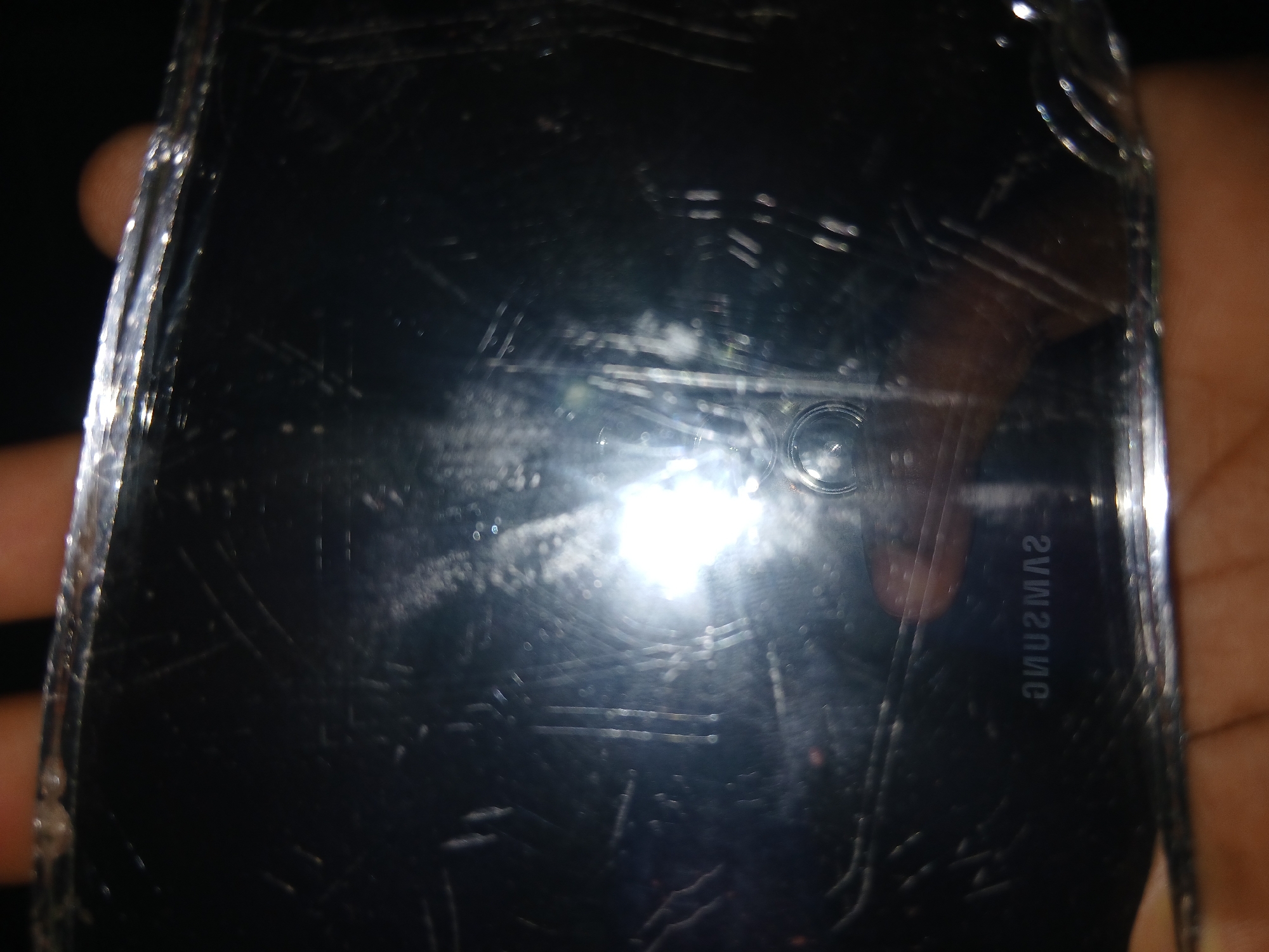 The image features a close-up view of a heavily scratched Samsung smartphone, held by a person. The phone's screen is black, indicating it is powered off or in standby. A bright, intense glare from a light source reflects prominently off the center of the screen, creating a significant hotspot that obscures the underlying details.
The screen surface is extensively damaged, covered with numerous visible scratches of varying depths, as well as apparent dust or smudges. Parts of a human hand, with light brown skin, are visible gripping the device along the left and right edges of the frame.
A distinctive detail is the mirrored text "SAMSUNG" clearly visible in the lower right section of the screen, confirming the brand of the device. In the upper right quadrant of the screen, a circular element, likely the front-facing camera lens or a sensor, can be discerned.
The background is dark and out of focus, suggesting an indoor setting with low ambient light, where the bright reflection off the phone screen is a dominant feature. The image primarily documents the physical condition of the phone's screen. While the location context is Lagos, Nigeria, no specific visual cues within the image itself confirm this. No particular activity beyond holding the phone is depicted, and no emotions are discernible.