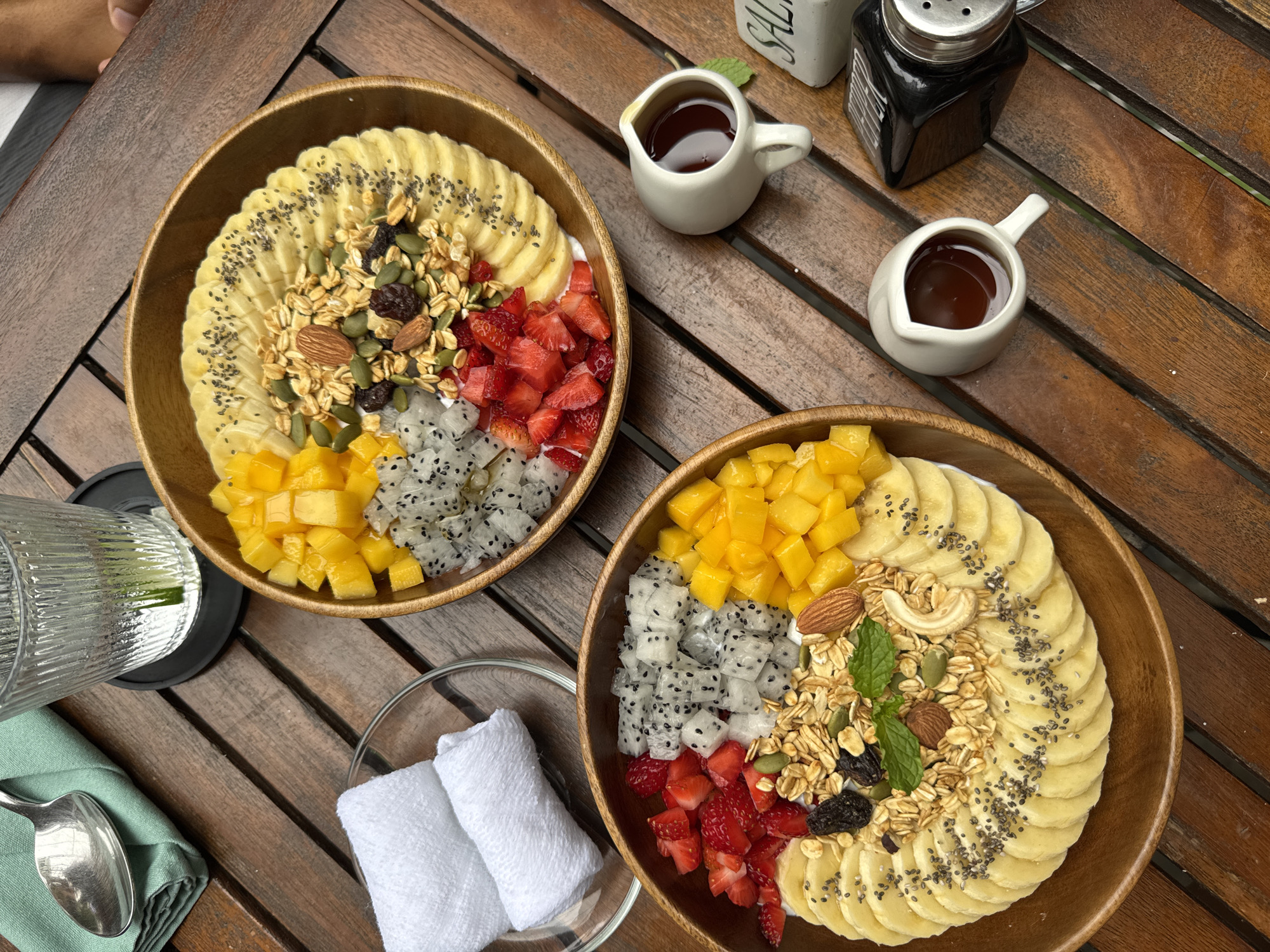 Two wooden bowls of fruit and granola are positioned on a wooden table. Each bowl contains sliced bananas topped with chia seeds, granola, pumpkin seeds, almonds, raisins, diced strawberries, diced mango, and diced dragon fruit over a base of yogurt. Two small pitchers of dark syrup sit nearby, along with a salt and pepper shaker. A glass with water, a spoon, and two rolled white towels sit on a clear plate on the left side of the frame. The scene appears to be photographed at an outdoor dining venue. While no specific landmarks are visible, the style and presentation suggest a location in a tourist area in the city of Đà Nẵng, Vietnam. No people are clearly visible in the frame, although there is a partial arm visible at the very top of the frame.