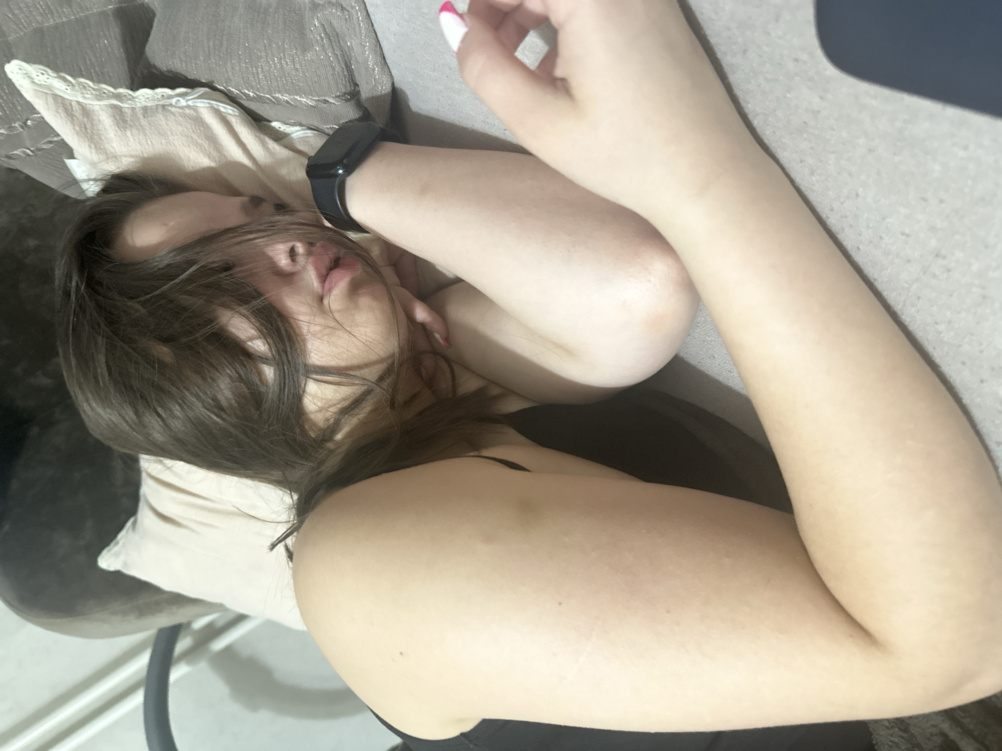 A young woman with dark hair is lying on her side on a couch. Her eyes are closed, and her head is resting on a pillow. She is wearing a black top and a black smartwatch on her left wrist. Her right arm is bent at the elbow, and her hand is resting near her face. Her left arm is extended along the couch. There are some faint marks on her left arm. The environment appears to be an indoor setting, possibly a living room. No specific location within Chișinău, Moldova can be determined from the image.