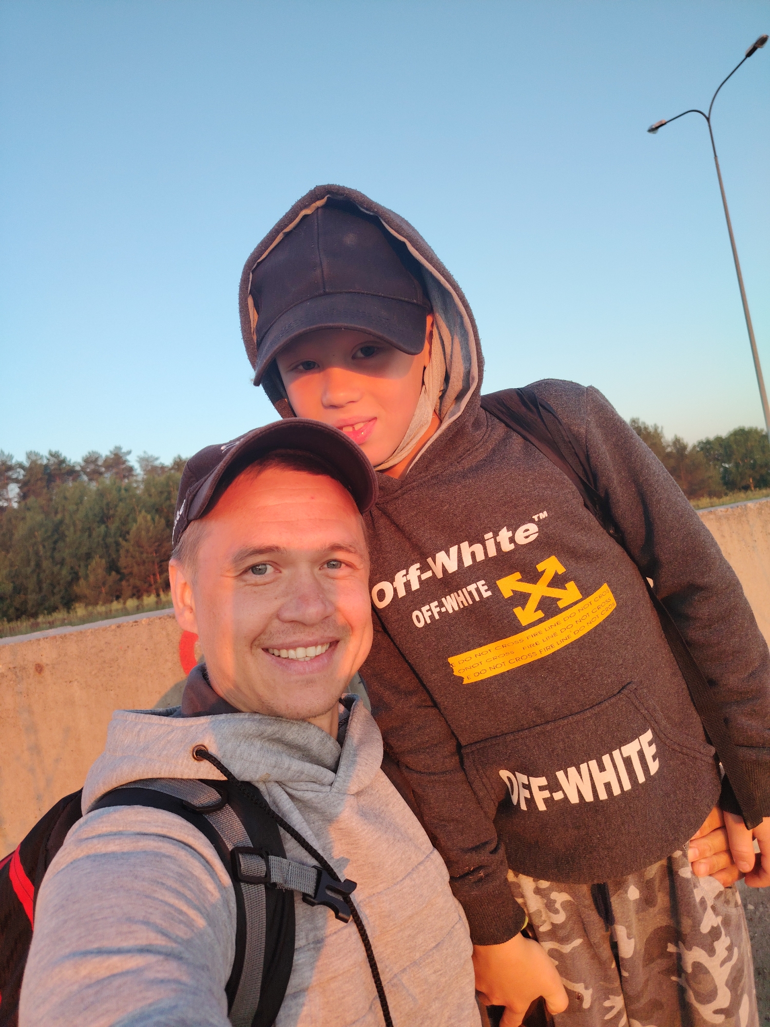 In the foreground, two individuals, an adult male and a youth, pose for a selfie against a backdrop of trees and a clear sky. The adult male, on the left, is wearing a grey hoodie and a black baseball cap. He is smiling broadly at the camera. To his right, the youth is wearing a dark grey hoodie with the "Off-White" logo and a black baseball cap. They are standing on a concrete barrier. The background reveals a line of trees and a street lamp against a pale blue sky, suggesting an outdoor urban or suburban setting, possibly on the outskirts of Chaikovsky, Russia, given the environmental cues. The lighting indicates it is either sunrise or sunset, casting warm tones on the subjects.
