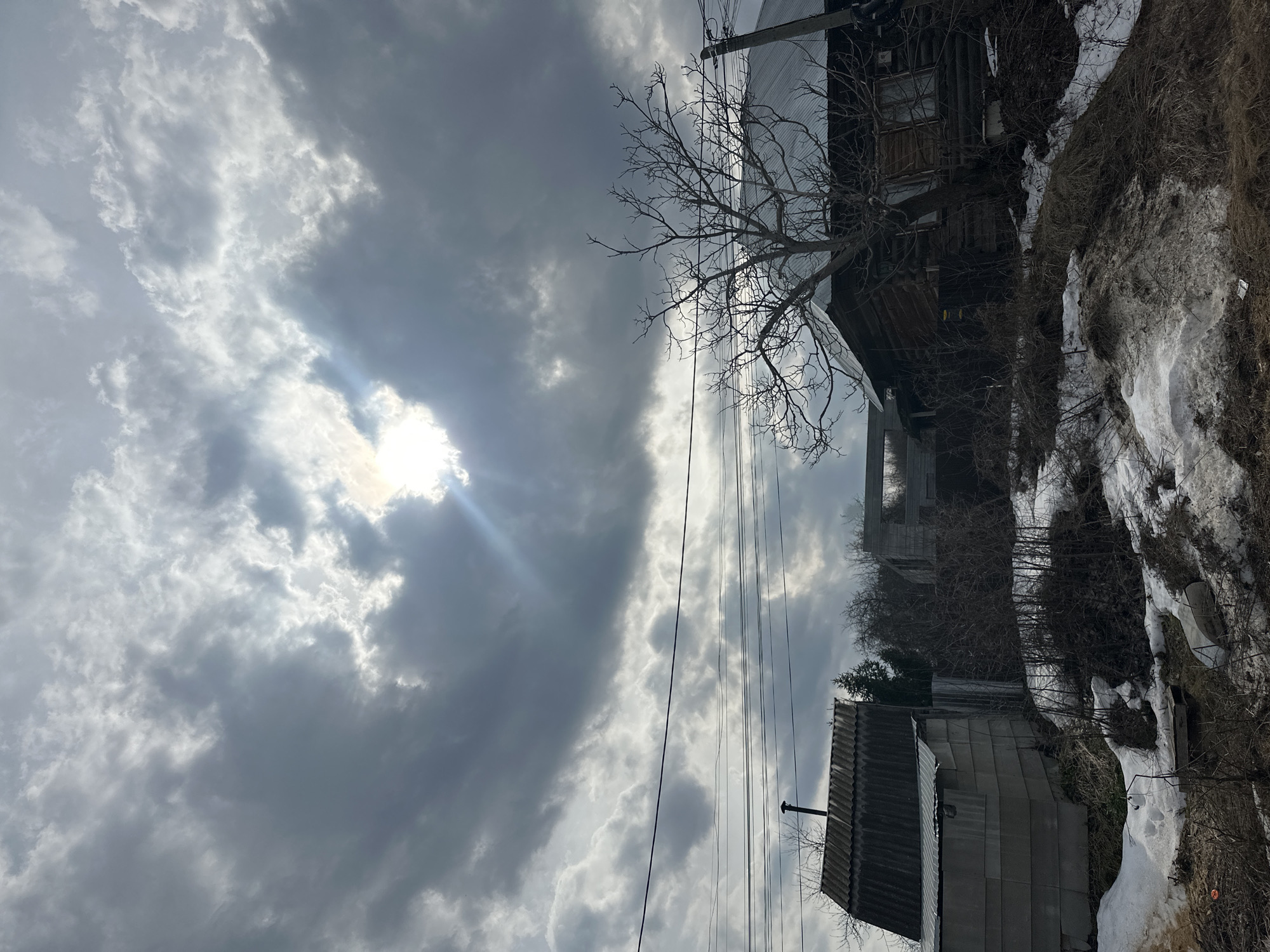 The image depicts a cloudy sky over a residential area in Chaikovsky, Russia. Sunlight breaks through the clouds. In the foreground, there is a gray metal building with a chimney. Above it, there are bare trees and wooden houses on a slope, with patches of snow visible on the ground. Utility poles and wires are present, crossing the sky.