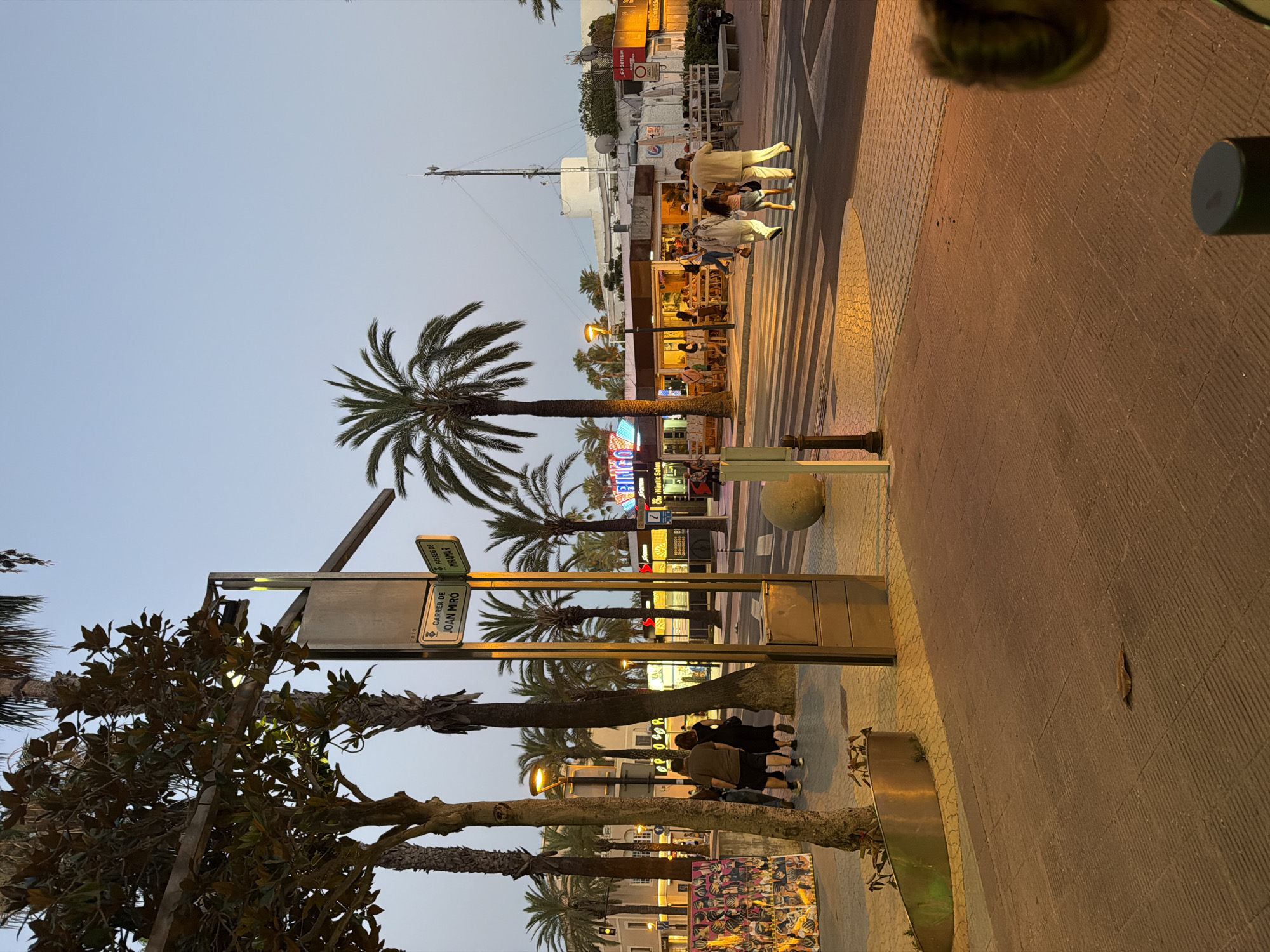 The image depicts a street scene in Salou, Spain, at dusk. A pedestrian crossing is visible in the foreground, with several people crossing. To the left, a street sign indicates "Carrer de Joan Miró" and "Passeig de Miramar." Palm trees line the street, and buildings with illuminated signs, including one for "BINGO," are present in the background. The sky is a pale blue, suggesting the transition from day to night. The overall atmosphere is that of a lively urban environment.