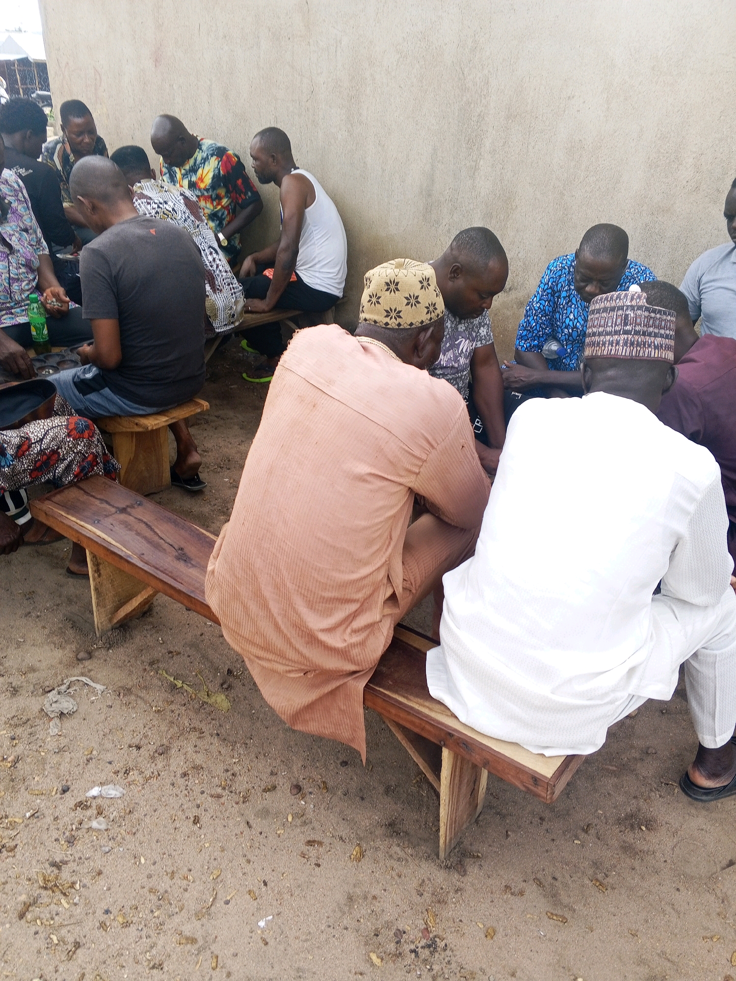 The image captures a gathering of men seated on wooden benches outdoors. The scene appears to be in Zariagi, Nigeria, with a textured, light-colored wall serving as a backdrop and sandy, uneven ground beneath the benches. Several men, mostly with dark skin and short hair, are visible, some wearing traditional African attire and head coverings, while others are in casual clothing.
The men are engaged in a group activity, with many of them looking down towards a central point, suggesting a shared focus on a game or discussion. One man in the foreground, wearing a white robe and a patterned cap, is the most prominent figure. To his left, another man in a peach-colored garment is seated. Further back, a group of men are clustered together, with one wearing a vibrant, multi-colored shirt.
The lighting suggests it is daytime, likely under overcast conditions or possibly during a late afternoon when the sun is not intensely bright. The environment appears somewhat rustic, with scattered debris on the ground, hinting at a less formal setting. The overall mood seems relaxed and communal, as the individuals share a common space and activity. No text is visible in the image.