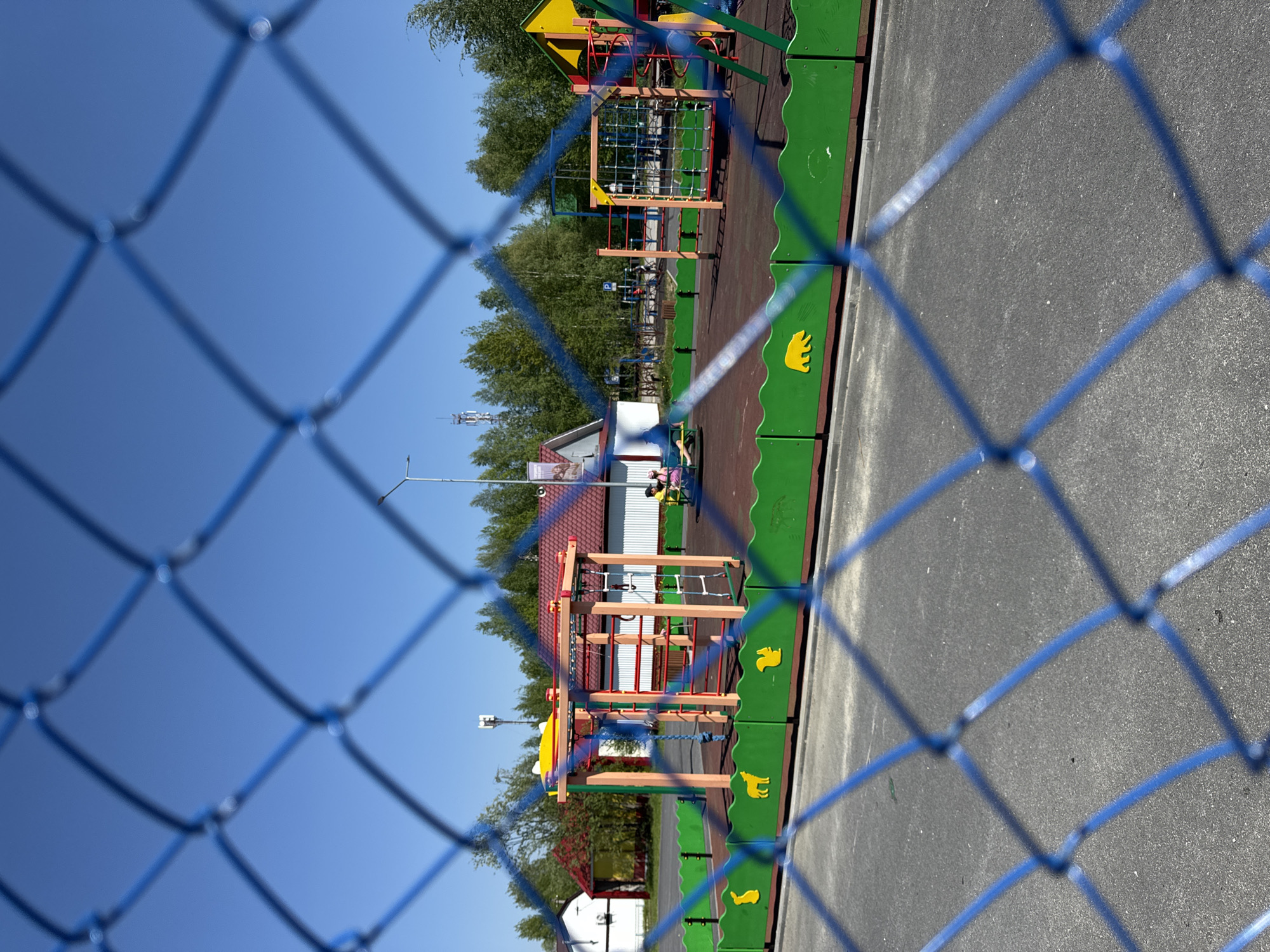 The image captures a vibrant children's playground in Высокий, Russia, viewed through a blurred, blue chain-link fence in the foreground. The fence forms a crisscross pattern, partially obscuring the scene beyond.
The playground features multiple colorful structures with wooden, yellow, and red elements, including climbing nets and ladders, set on a dark reddish-brown soft-fall safety surface. A distinctive green barrier, adorned with yellow animal cutouts such as a bear, squirrel, and possibly a fox or dog, lines the perimeter of the play area on the left side. To the right of the playground, a grey asphalt or paved surface is visible.
In the background, a small white building with a red-tiled roof stands to the left. Further back, dense green trees frame the scene under a clear, bright blue sky. A tall pole equipped with multiple antennas is discernible among the trees, suggesting a modern infrastructure element. Indistinct figures, likely people, are visible near the playground equipment and the white building, hinting at ongoing activity or recent use.
The scene is illuminated by bright sunlight, indicating a clear, sunny daytime. No specific activities are clearly defined, but the setting suggests a lively recreational space. While there are signs on distant buildings, no text is legible. The overall impression is one of an inviting outdoor play area on a pleasant day.