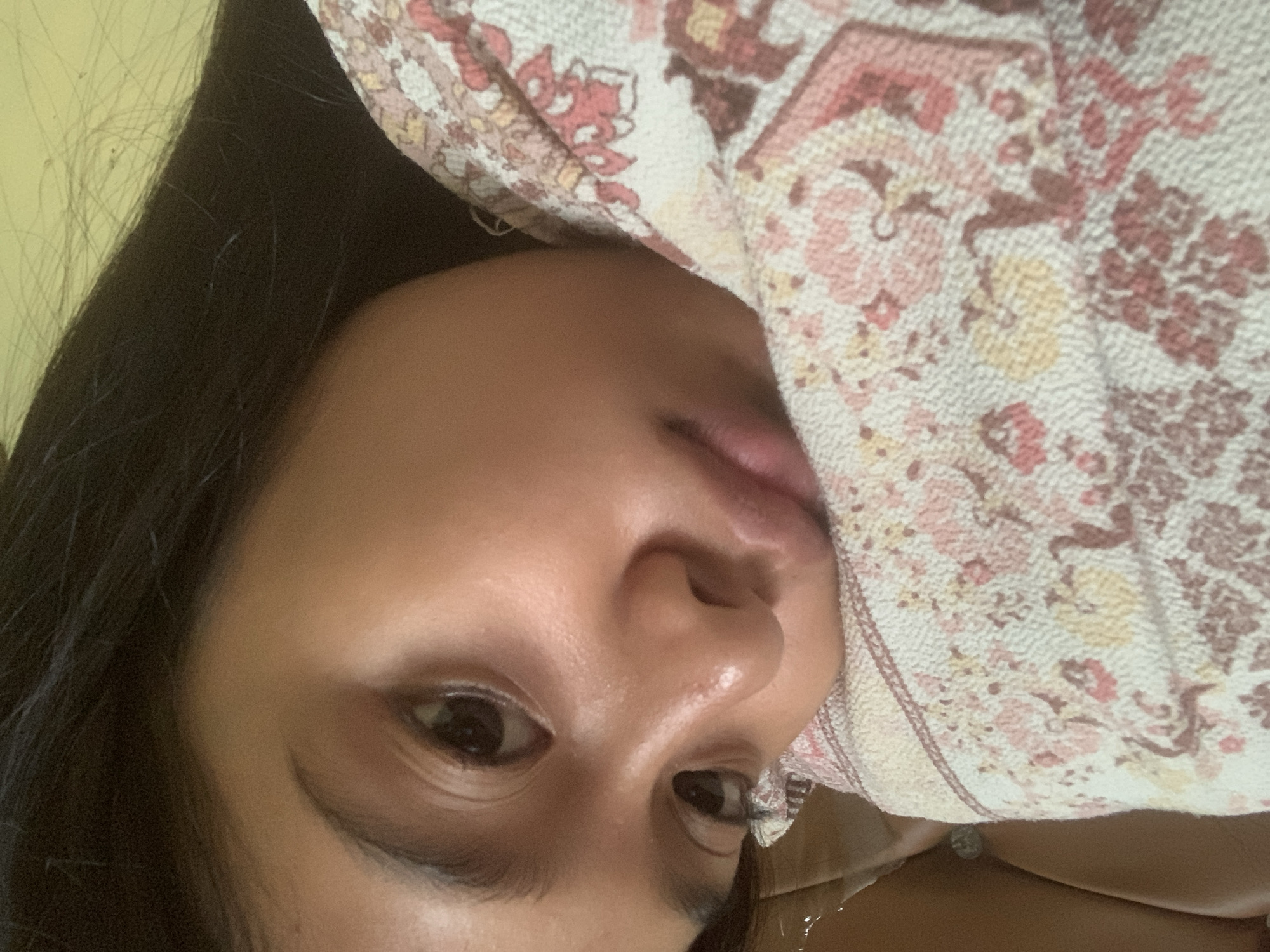 A close-up, low-angle selfie of a young person is shown, featuring their face and upper body. The subject's head is tilted to the left, with their right eye partially visible and their left eye prominently displayed. Their dark hair frames their face. The individual is resting their cheek on a blanket with a floral pattern in shades of pink, red, and yellow. The background consists of a plain, light-colored surface, possibly a wall. The image appears to be taken indoors, with soft lighting. No specific location in Sungai Meriam, Indonesia, is depicted or suggested by the visual information in the image.