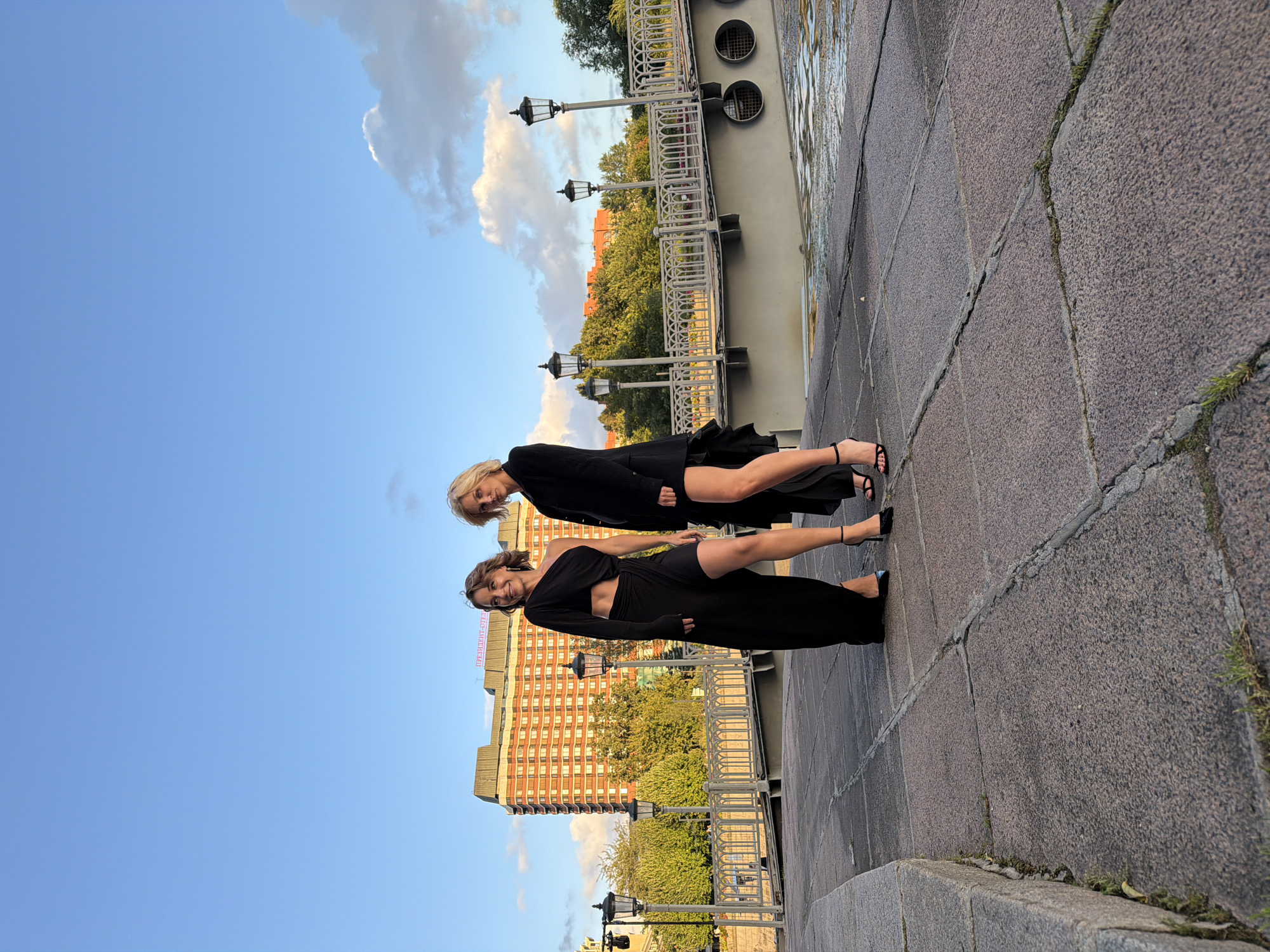 Two women are standing on a paved walkway near a body of water, with a bridge and buildings in the background. The woman on the left is wearing a black one-shoulder dress with a high slit and black high heels. The woman on the right is wearing a black suit jacket with a short skirt and black high heels. The building in the background is red brick with many windows and has a sign that reads "ПРЕЗИДЕНТ-ОТЕЛЬ" which translates to "President Hotel". The bridge has decorative railings and lampposts. The sky is blue with scattered clouds. This appears to be a public space in Moscow, Russia, likely along the Moskva River, given the presence of the President Hotel.
