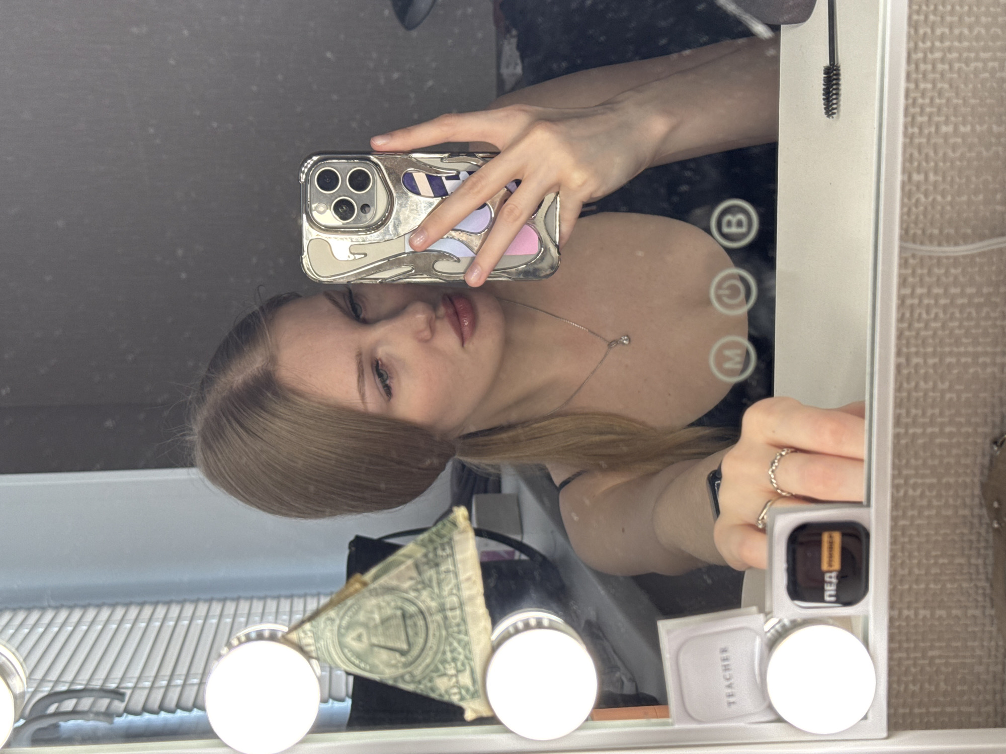 A young woman is taking a selfie in a mirror with lights. She is holding a cell phone with a decorative case in her right hand. She is wearing a necklace. Her hair is light brown and pulled back in a low ponytail. A one dollar bill is wedged between lightbulbs at the bottom of the mirror. Cosmetic items are also visible at the bottom of the mirror. The image was likely taken indoors. There is no information to suggest this photo was taken in Perm, Russia.