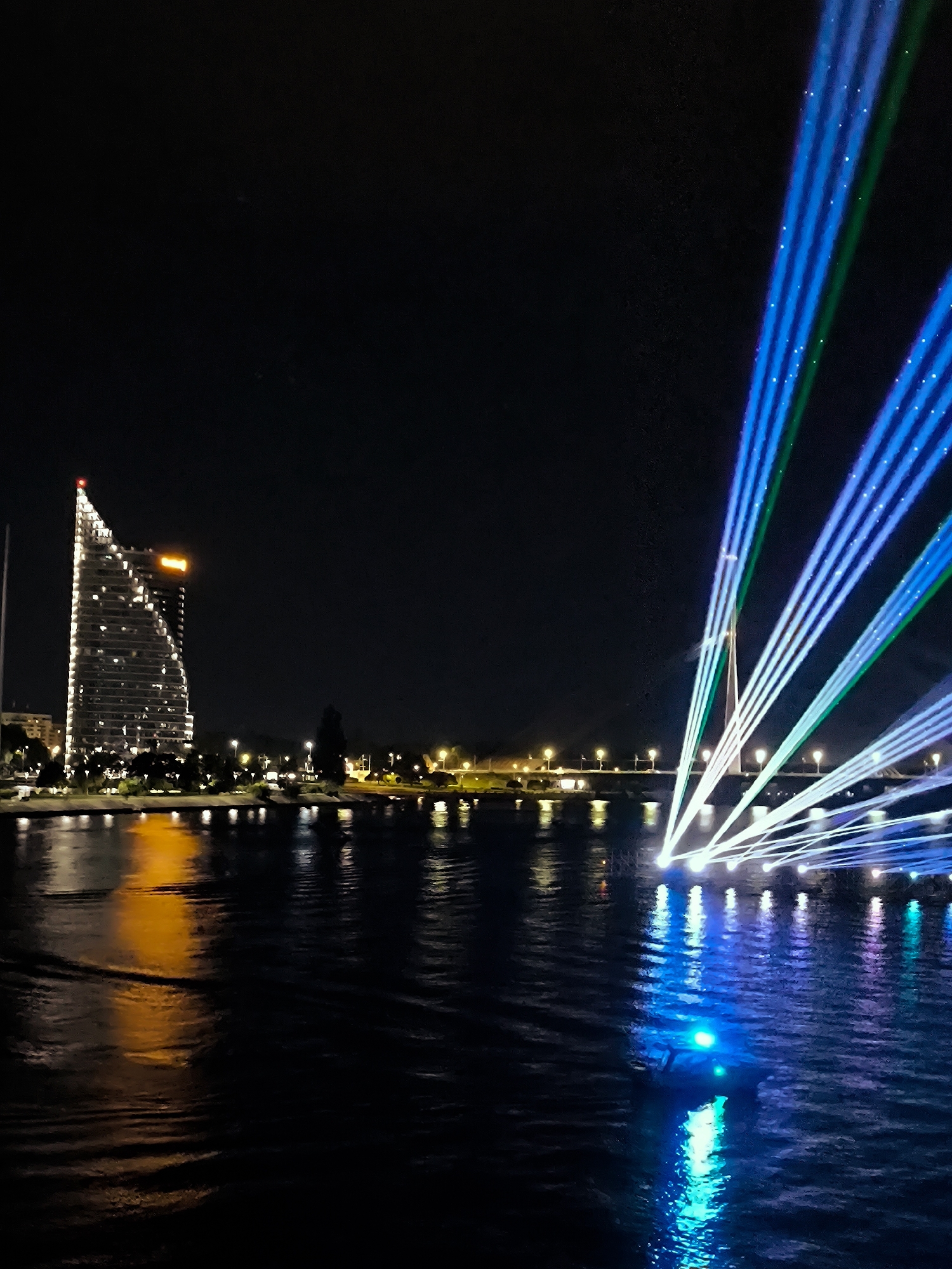 The image captures a nighttime scene on the Daugava River in Riga, Latvia. A modern, illuminated skyscraper, likely the Z Towers, dominates the left side of the frame, its glass facade reflecting the city lights. The river in the foreground shows reflections of the building and the vibrant, multicolored laser light show projected across the water from right to left. The laser beams, a mix of blues, greens, and whites, create a striking visual spectacle against the dark sky and water. A bridge structure is faintly visible in the background, with lights lining its span. The overall impression is one of a lively urban environment illuminated by impressive light art. There are no people or animals clearly visible in the image.