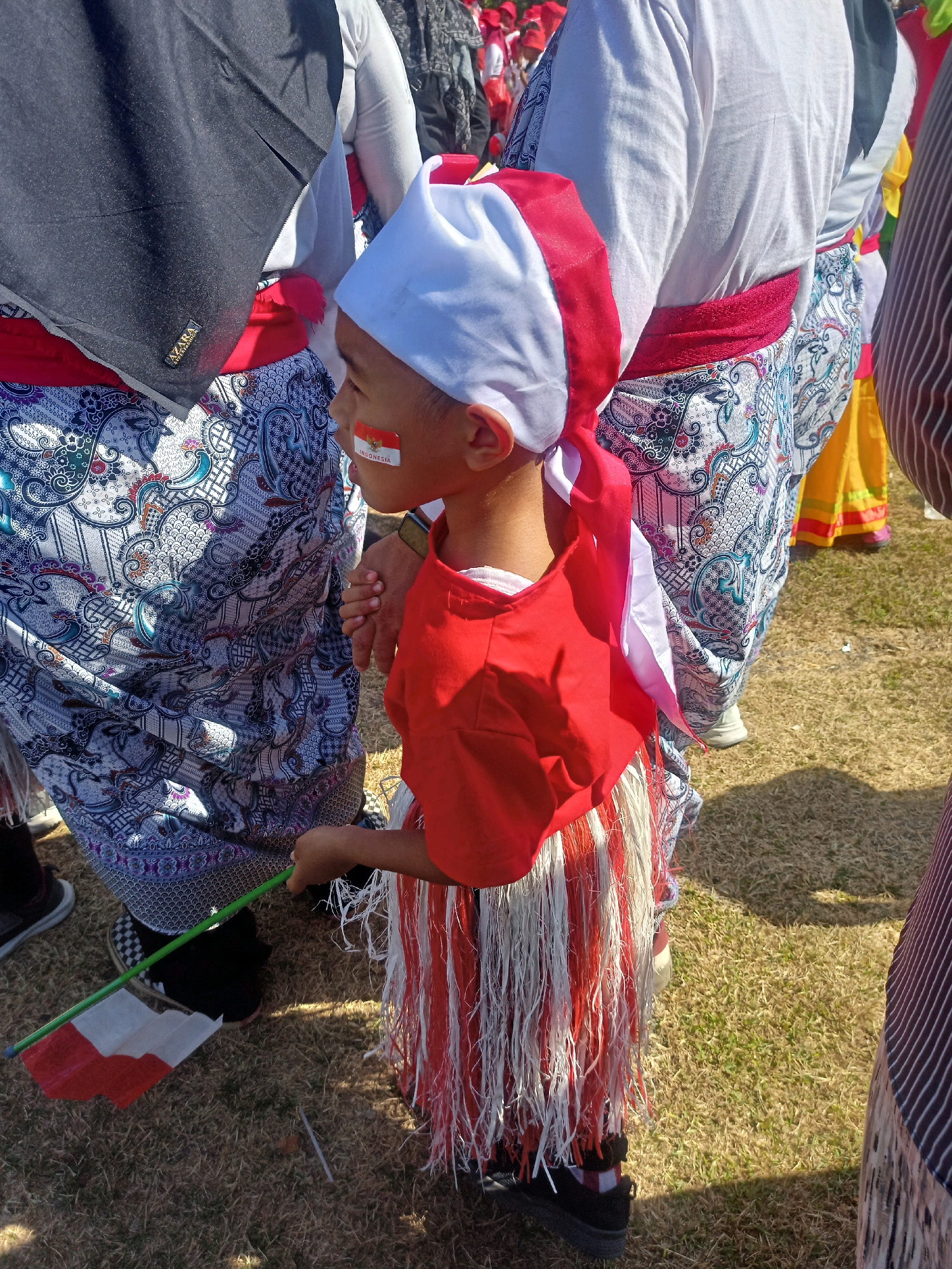 The image captures a young boy dressed in traditional Indonesian attire, adorned with red and white colors. He wears a red shirt and a patterned skirt with white and red fringe. A red and white bandana covers his head, and the Indonesian flag is painted on his cheek. He holds a green stick with a small Indonesian flag attached. He stands on dry grass, with other people in similar traditional clothing around him. One person on his left wears a black hijab, and another person on his right is partially visible with a striped fabric. The scene appears to be outdoors, likely during a cultural event or celebration in the city of Sumberlawang, Indonesia.
