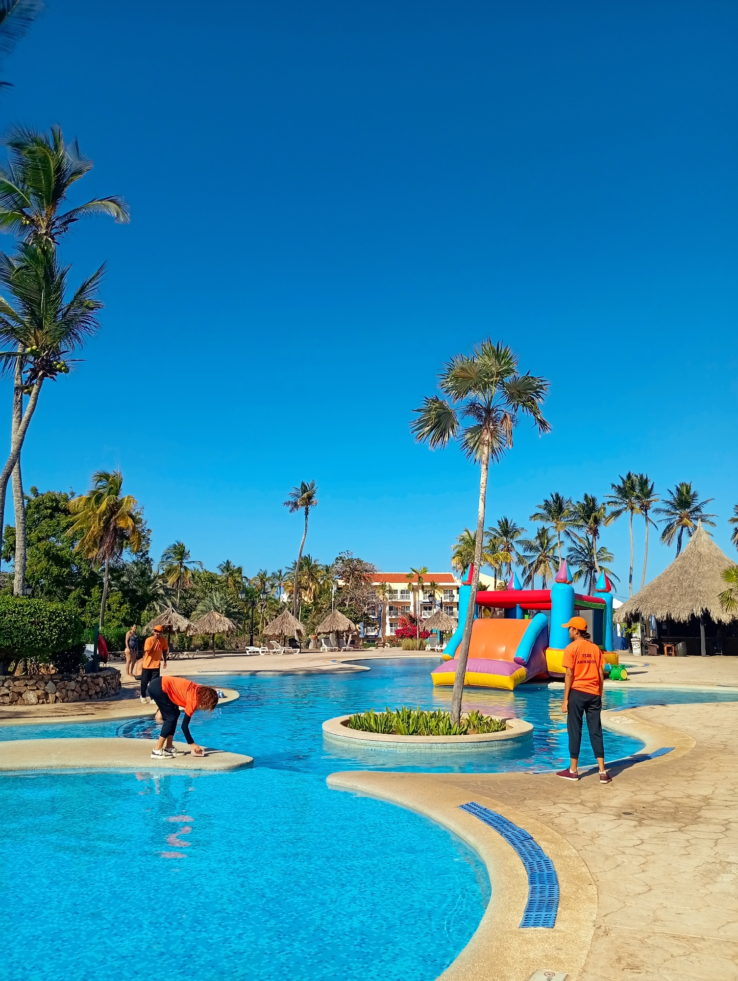 The image shows a sunny, tropical resort scene likely in Playa El Agua, Venezuela, given its beach setting and resort architecture. A blue-tiled pool dominates the foreground with two employees wearing orange "Team Animation" shirts present: one bending over on a small island in the pool and another standing near the edge. A colorful inflatable slide is positioned near the pool, suggesting recreational activities. Palm trees are scattered throughout the scene, and thatched-roof huts are visible in the background. The sky is clear and blue. In the background, a resort building can be seen.
