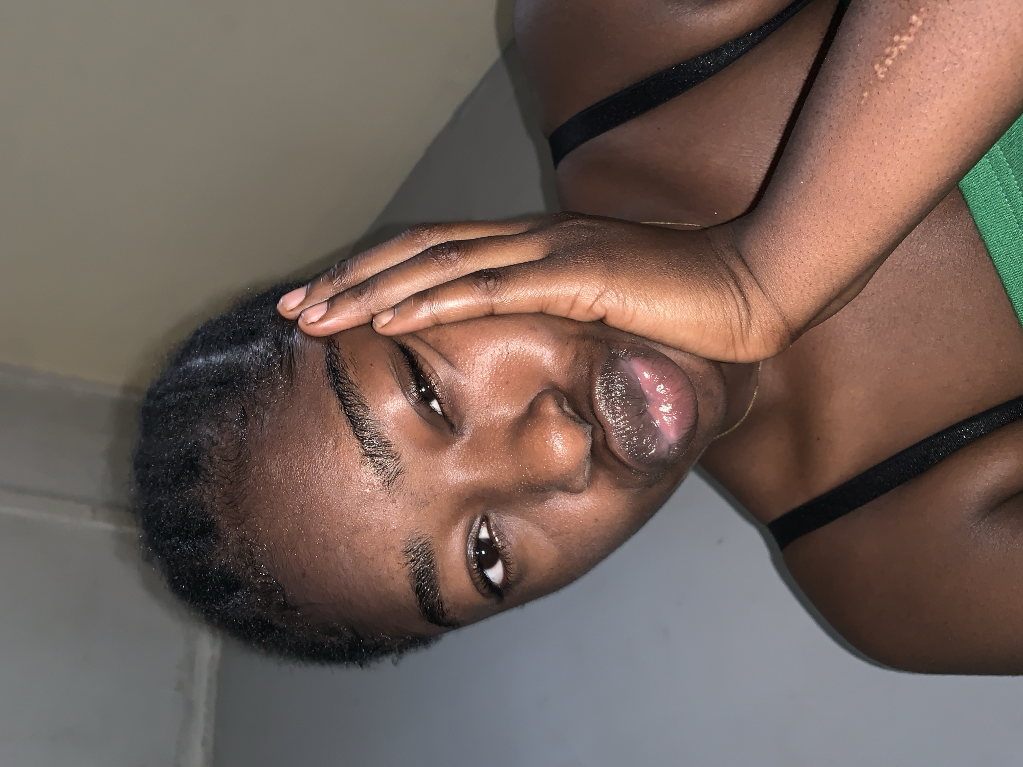 A close-up portrait of a dark-skinned woman with short, dark hair. She is wearing a green top with black straps and facing the camera with her right hand on her forehead. Her lips appear to be wet or glossy. The background shows a wall with an undefined light source. The photo does not contain sufficient information to confirm the location as Rafin Par, Nigeria. The image presents only the subject's face and partial upper body, and details about the environment.