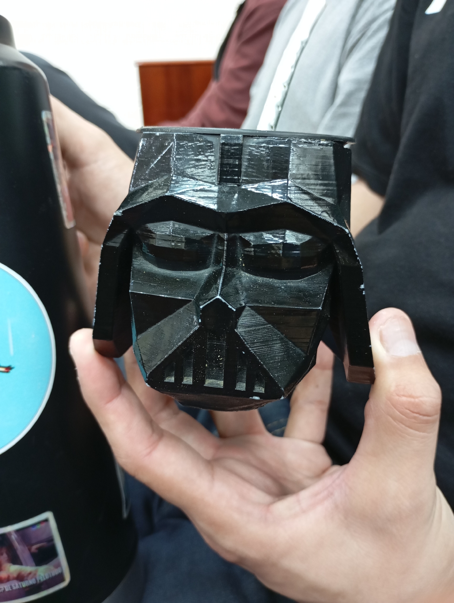 The image shows a close-up of a person holding a black, 3D-printed Darth Vader mask. The mask is detailed, with a faceted design and visible layer lines from the 3D printing process. The person's hands are visible, supporting the mask from underneath and on the side. In the background, other individuals are partially visible, wearing casual clothing. To the left of the mask, a large black object with a light blue circular sticker is present, partially obscuring a sticker at the bottom which appears to have text in Spanish. The scene appears to be indoors, with a blurred background suggesting a casual gathering or event. There is no specific visual information within the image that definitively places the scene in La Plata, Argentina.