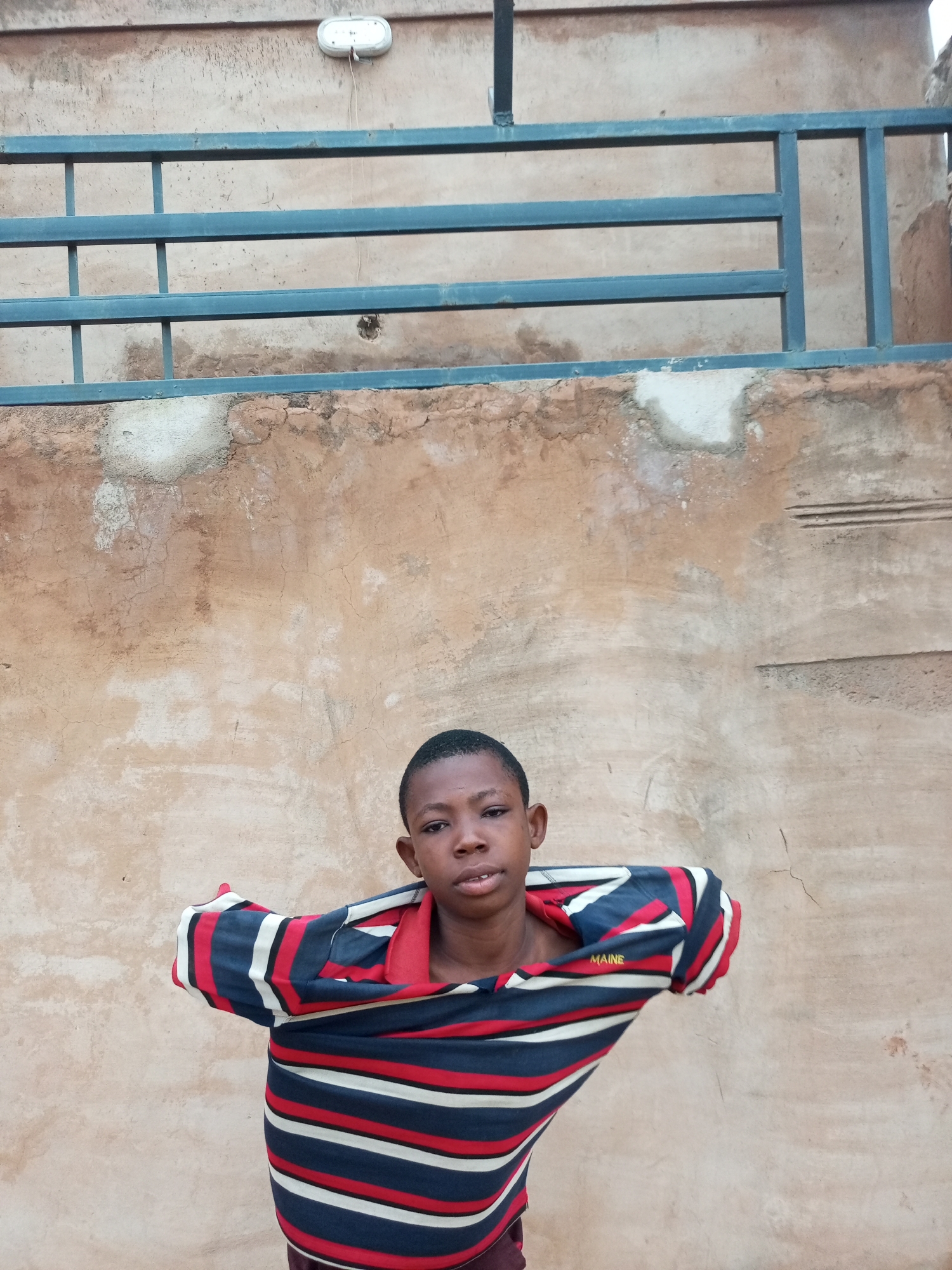 A young Black boy with short hair, wearing a horizontally striped t-shirt with navy, red, and white bands, is standing in front of a textured, light brown wall. His t-shirt is partially off, with one sleeve pulled up and the other hanging loose. The wall has areas of peeling plaster and what appear to be faint scratches or markings. Above the wall, a blue metal railing is visible, with horizontal bars and vertical supports. At the top of the image, near the center, a white oval light fixture is mounted to the wall. The scene appears to be outdoors, possibly in an urban setting in Tempure, Nigeria.