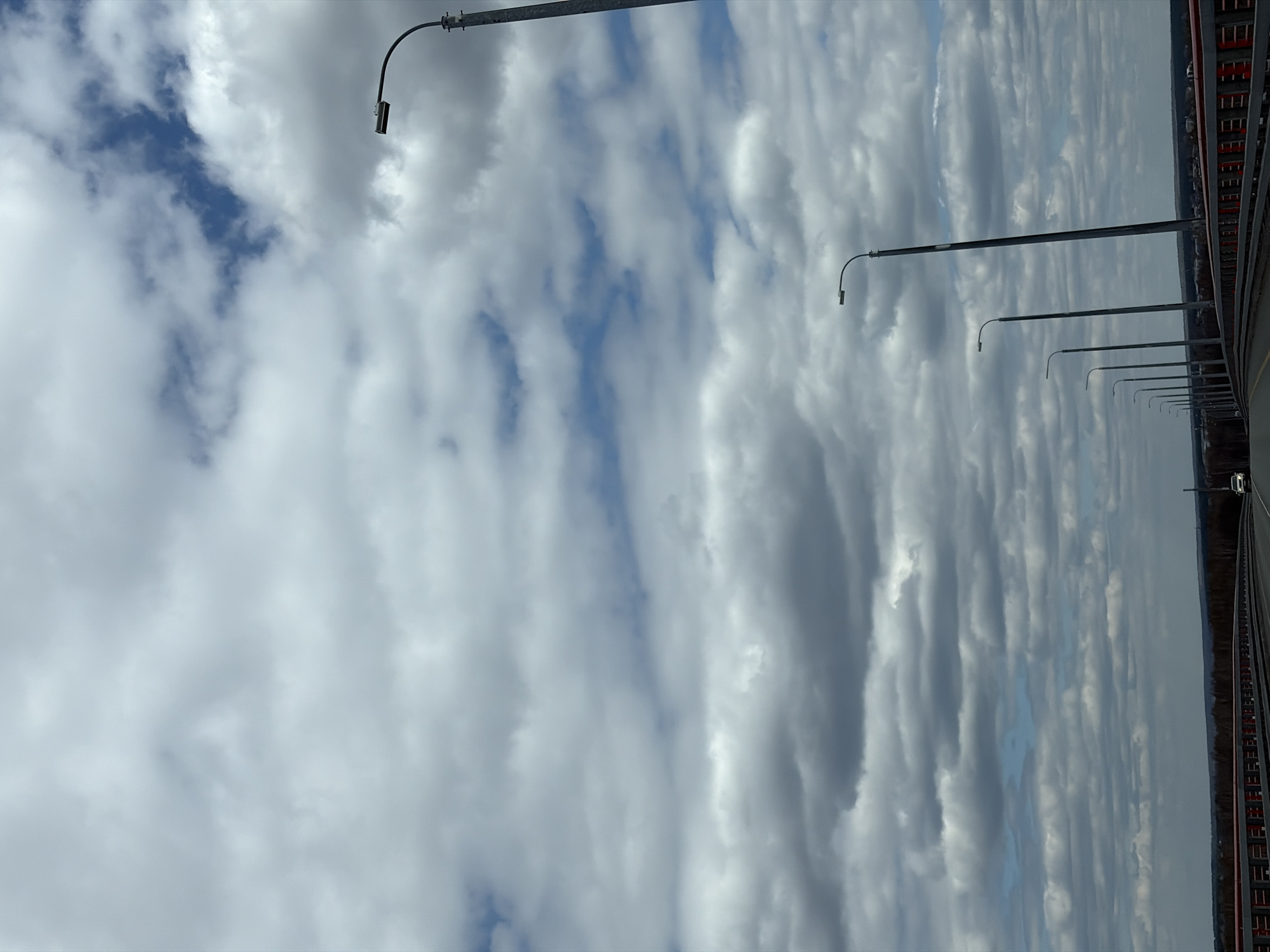 The image shows a view from a bridge in Tarasovo, Russia. The sky is filled with layered, striated clouds with patches of blue visible. A row of streetlights runs along the right side of the frame, attached to the bridge's railing. The bridge's structure, including a red metal railing and potentially other structural elements, is partially visible on the far right. The general environmental conditions indicate an overcast or partly cloudy day. A road and maybe a car are visible on the bridge.