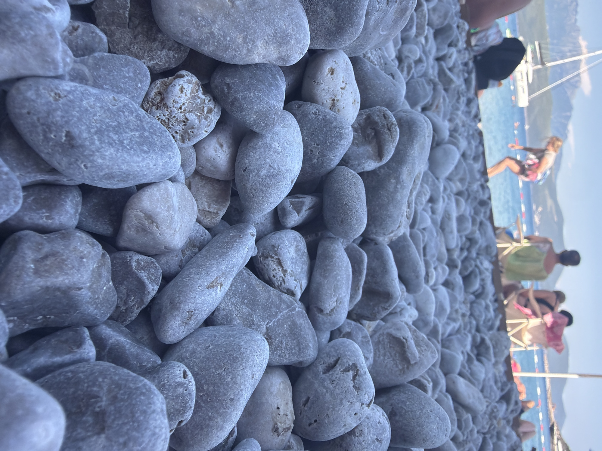 This is a vertical, close-up, eye-level shot of a pebble beach. The foreground is dominated by an abundance of smooth, grey and white pebbles of various sizes and shapes, with some larger, oval-shaped stones being most prominent. The texture of the pebbles is detailed, showing some porous surfaces and natural variations in color and pattern.
In the background, the scene transitions to a blurred view of a beach resort area. The turquoise water of the sea is visible, with some boats and a sailboat on the horizon. There are also glimpses of people on what appears to be a pier or boardwalk. One person is clearly visible, wearing a bathing suit and seemingly in motion, possibly engaging in a water sport. Other figures are also present, some with towels or beachwear. The overall atmosphere suggests a sunny day at a popular tourist destination.
The time of day appears to be late afternoon or early evening, given the warm, soft lighting and the long shadows that might be implied by the scene. The weather seems pleasant and clear, suitable for beach activities. The location context provided, Radovići, Montenegro, further reinforces the image's setting as a coastal area in the Mediterranean. No visible text is present in the image. The image captures a sense of relaxation and summer vacation vibes.