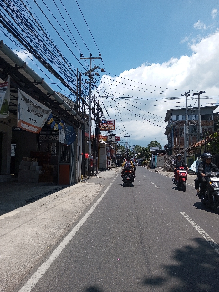 This image depicts a street scene in Tagtse Tashidingkha, Bhutan, under a clear blue sky with scattered white cumulus clouds. The view is down a paved asphalt road, which features a solid white line marking its right edge. Along the left side of the road, a concrete sidewalk runs parallel to a series of commercial buildings. The nearest building on the left exhibits a banner with text reading "TOKO BANGUNAN DAN ALAT LISTRIK" and "TS METRO" in Indonesian, indicating a building materials and electrical tools shop. Stacks of light-colored bricks or blocks are visible within this storefront. Numerous utility poles line both sides of the street, heavily laden with a dense, crisscrossing network of overhead electrical and communication wires. Three individuals are riding scooters on the road, traveling away from the viewer. In the foreground right, a person wearing a dark helmet and dark clothing operates a black scooter. In the mid-ground right, another person, also in a dark helmet and dark attire, rides a red scooter. Further ahead in the mid-ground center-left, a third individual wearing a yellow top, a light helmet or head covering, and a backpack, rides a dark scooter. Shadows from overhead structures and objects are cast upon the road surface. In the background, additional multi-story buildings, including a brick and concrete structure, and some trees are visible.