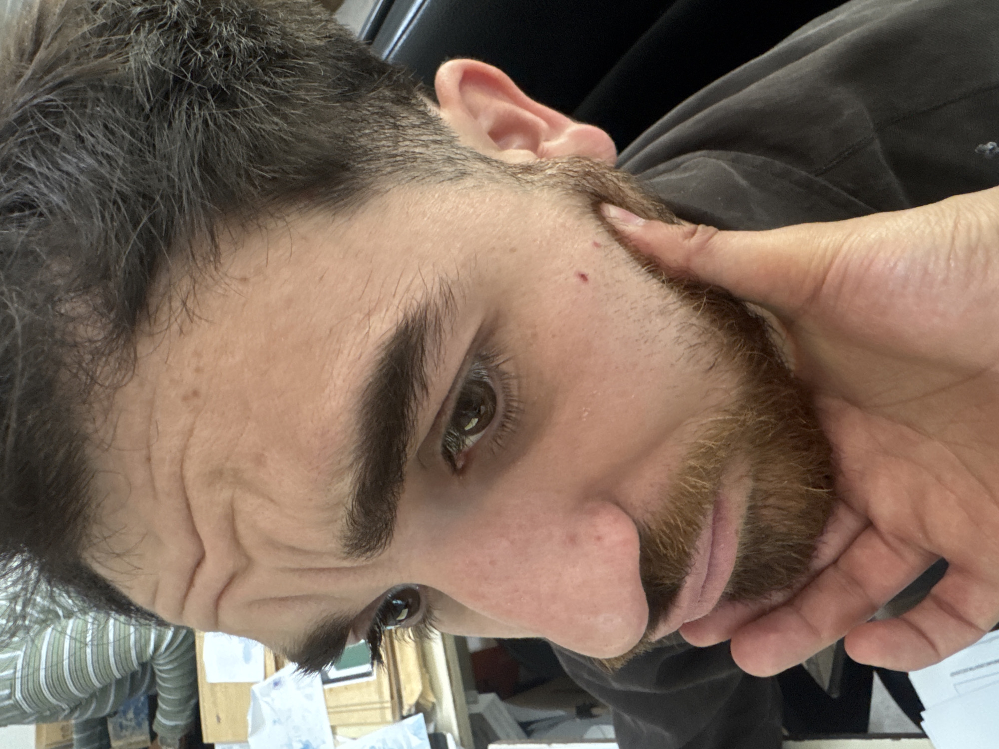 A close-up, high-angle portrait shot features a man in Puebla, Mexico, with his head tilted approximately 90 degrees to the right. He has dark, somewhat messy hair with visible lighter strands, and thick, dark eyebrows. His brown eyes are cast downwards, contributing to a pensive or thoughtful expression.
A reddish-brown beard and mustache frame his lower face. His left hand is raised, with fingers gently cupping or gripping his beard and jawline. Several horizontal wrinkles are visible on his forehead, accentuated by the head tilt and expression. Small reddish blemishes are noticeable on his cheek and the bridge of his nose.
The man is wearing a dark, long-sleeved shirt or jacket. The blurred background indicates an indoor setting, possibly an office or workshop, with a dark object behind his ear and faint glimpses of papers on a surface below. On the far left, a person or object in a light green and white striped garment is partially visible. No discernible text is present in the image.