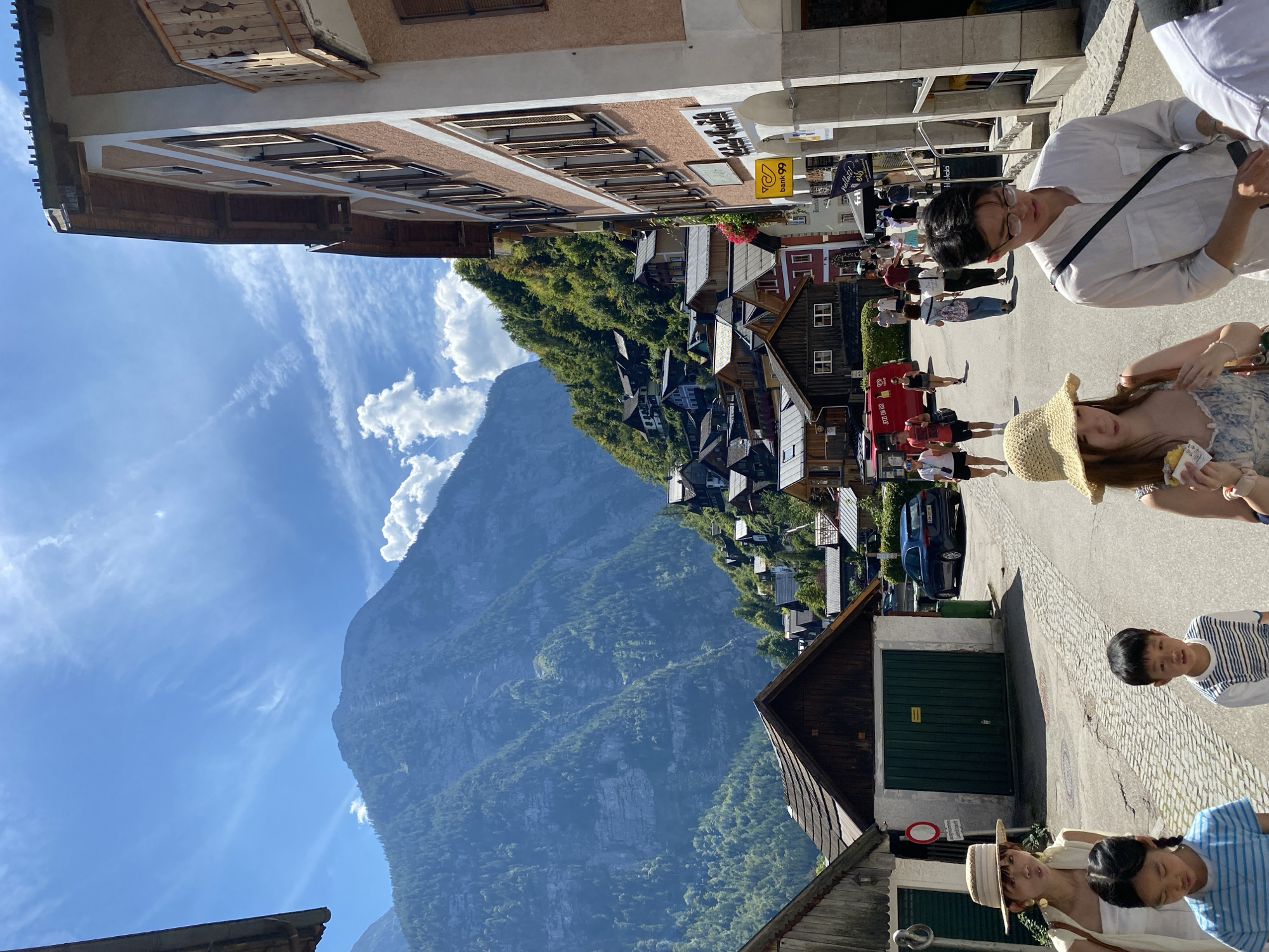 In Hallstatt, Austria, on a sunny day, a group of tourists walks down a cobblestone street. Mountains covered in lush green trees tower over the picturesque village. The buildings are a mix of traditional Austrian architecture with wooden balconies and pastel-colored facades. On the right side of the street, a building with a peach-colored exterior features multiple windows with wooden shutters and a small balcony. A yellow "Bank 99" sign is visible on the building. On the left side, a wooden structure with a dark roof and a green garage door stands. Several people are walking in the street, including a woman in a straw hat and a white top, two children in casual clothing, and a man wearing glasses and a white shirt. The sky is a vibrant blue with scattered white clouds.