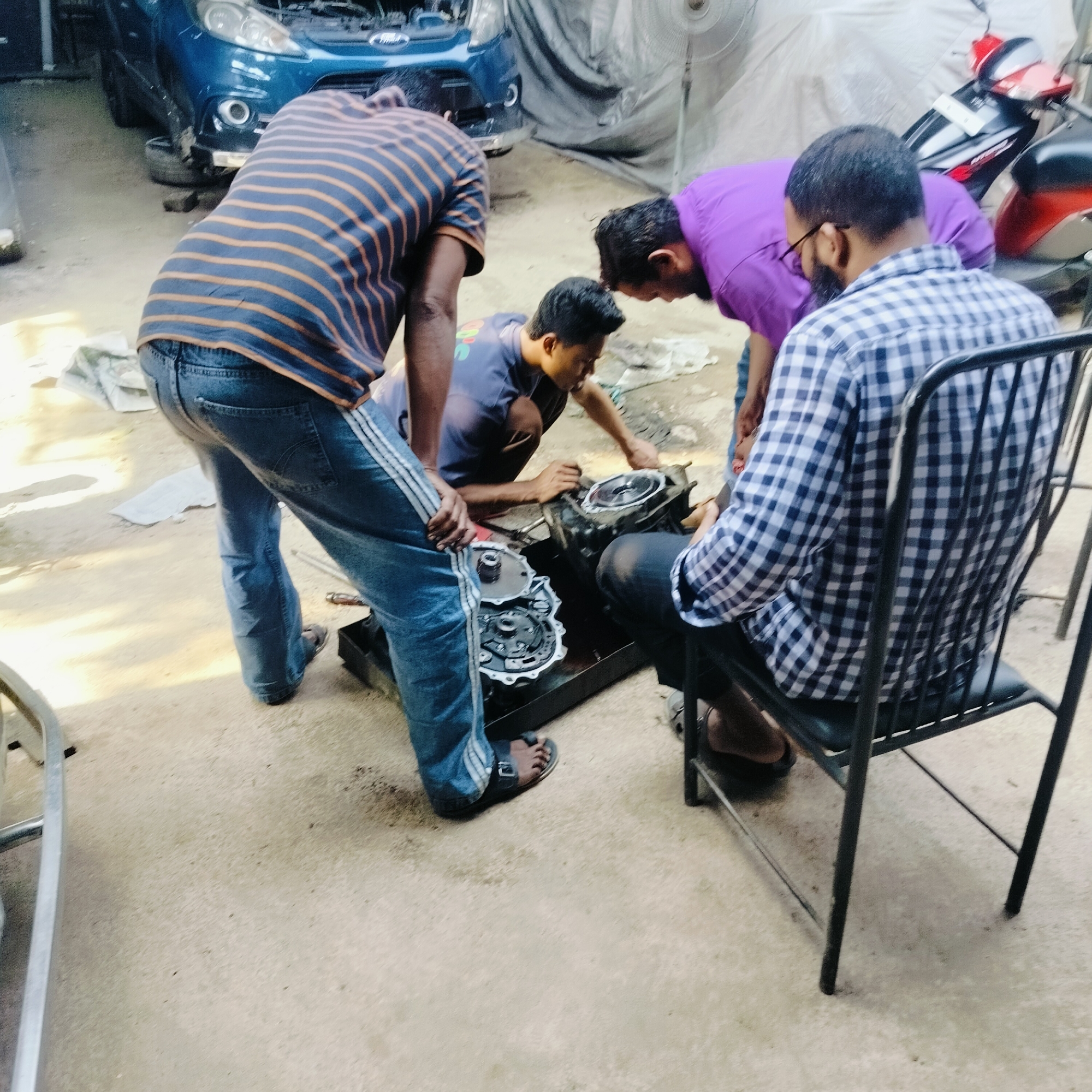 In an open-air auto repair shop in Dhaka, Bangladesh, four men are gathered around what appears to be a dismantled engine or transmission component. One man, wearing a horizontally striped shirt and jeans with white stripes down the side, leans over the machinery. Another man, wearing a purple shirt, is also leaning over, appearing to examine the parts. A third man, in a blue shirt, is kneeling and working directly on the engine component. The fourth man, wearing a blue and white checked shirt, is seated in a black chair, observing. Tools and parts are scattered around the work area. A blue car with its hood open is parked in the background, along with a red and black motorcycle. A white cloth or tarp is draped in the background, partially obscuring a fan. The floor of the shop is concrete.