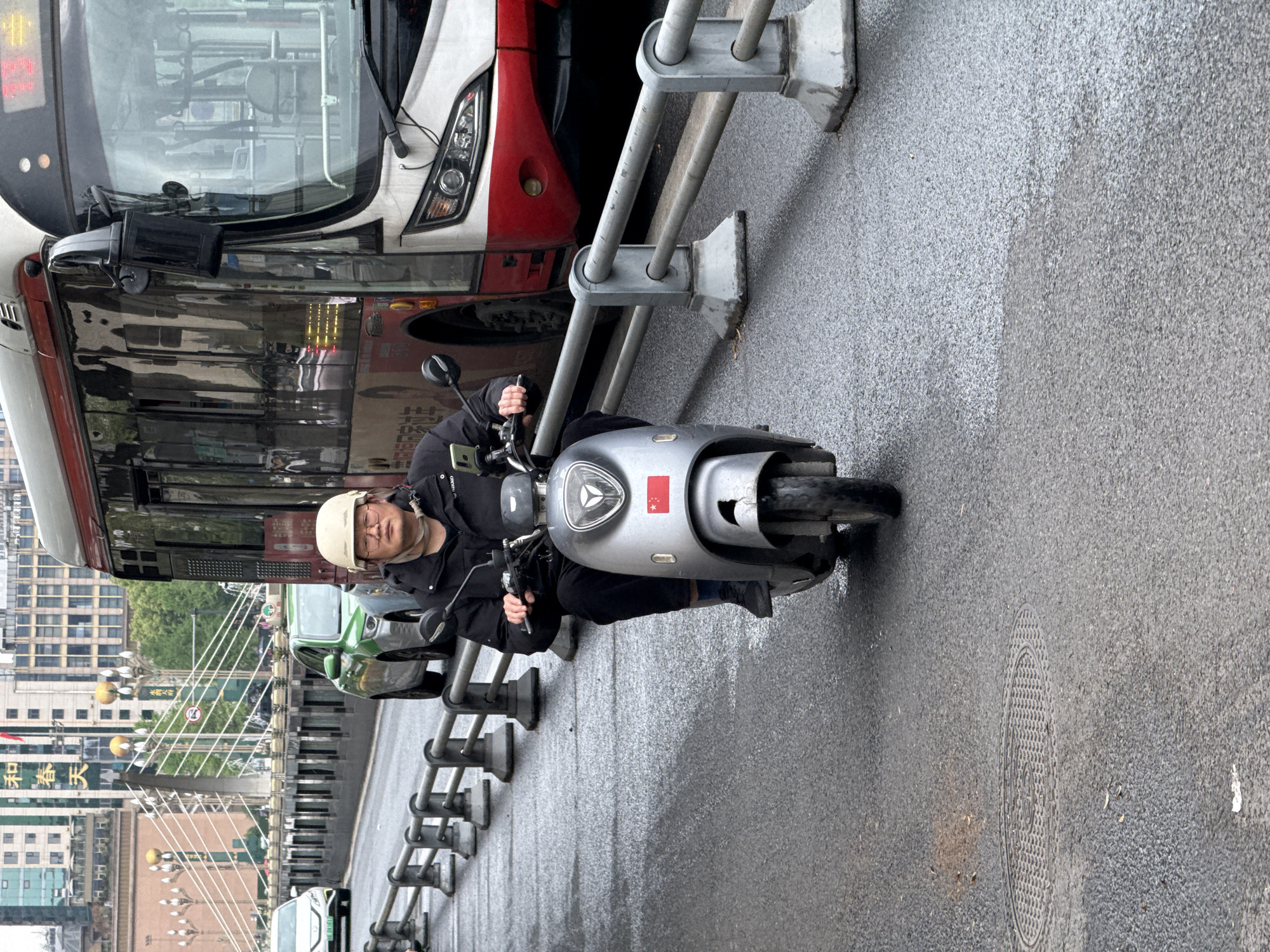 A man wearing a white helmet and glasses is riding a silver scooter on a wet, gray road next to a bus. A silver guardrail separates the scooter from the bus. The scooter has a Chinese flag sticker on the front. In the background, there's a cityscape with buildings, trees, and streetlights. A bridge is visible, with signs in Chinese characters. The location is identified as Qingyang District, China.