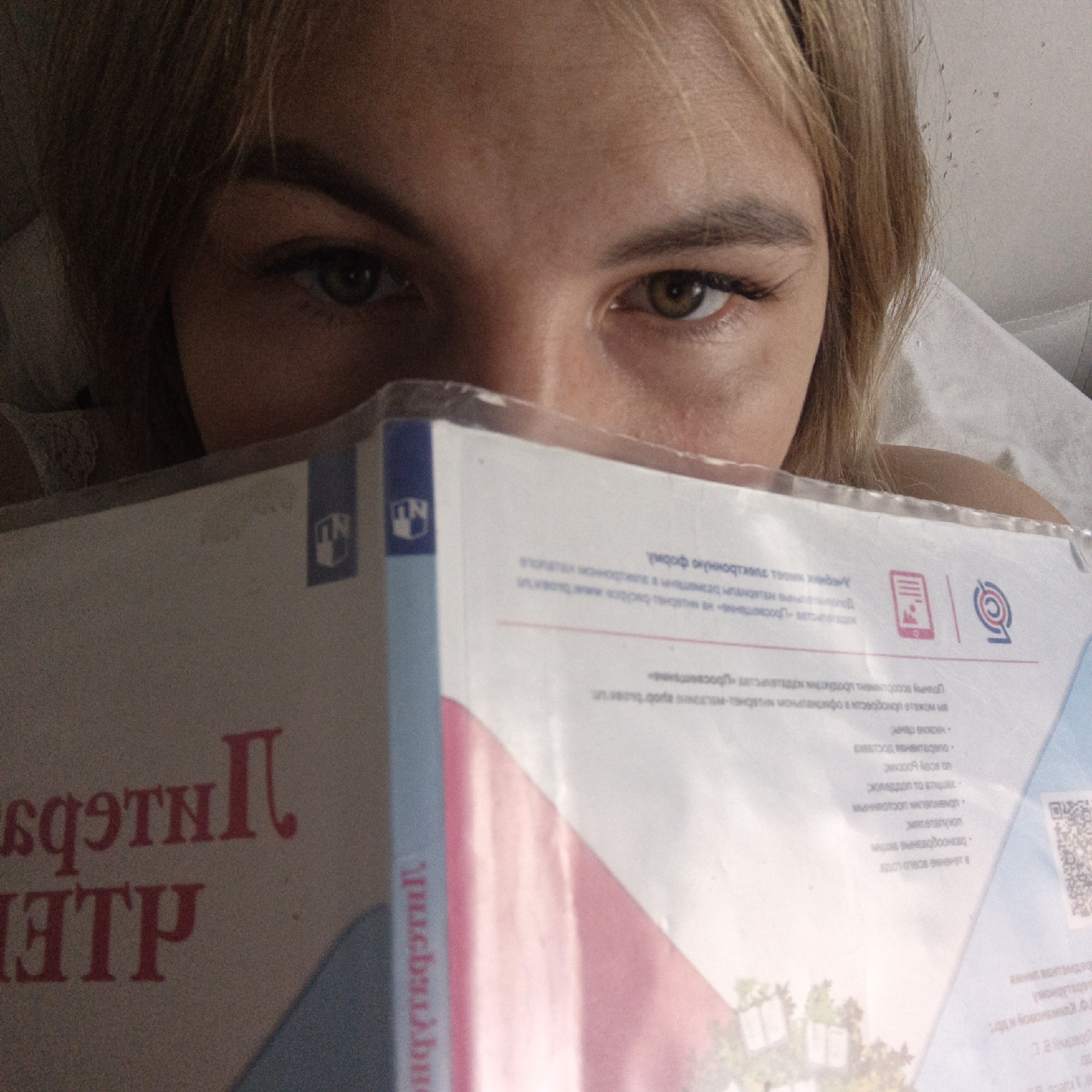 A close-up image shows a person, likely a woman, looking directly at the viewer over a document or book that partially obscures her face. Her light brown or blonde hair frames her face, and her prominent greenish-brown eyes are visible. The setting appears to be indoors, possibly a bedroom, with a soft, light-colored surface like a pillow or bedding visible in the background.
The person is engaged in an activity that involves holding and looking at the document. The document's cover is visible, featuring text in Cyrillic, likely Russian, and several logos.
Notable details from the visible text and logos include:
- On the left side of the document, mirrored red text reads: "ИНТЕЛЛЕКТ" (Intellect), "ПАРТНЕР" (Partner), and "ЦЕНТР" (Center). This suggests the document might be from an "Intellectual Partner Center" or similar organization.
- On the right side, non-mirrored text mentions terms like: "Фонду" (Fund), "программы" (programs), "мобильное приложение" (mobile application), "Учебные пособия" (Textbooks/Learning aids), "Яндекс.Кью" (Yandex.Q), "Сборник документов" (Collection of documents), and "Минобрнауки России" (Ministry of Education and Science of Russia).
- Logos include two instances of a stylized "ND" or "ПИ" (PI) within a shield shape, an icon of a mobile phone with a document, and a stylized 'Q' or target symbol, often associated with online platforms.
The overall context suggests the person is reading or studying educational materials, possibly related to a government educational program or an organization like Yandex.Q, with components involving mobile applications and digital resources. The lighting is soft and ambient, and the person's expression appears neutral and focused.