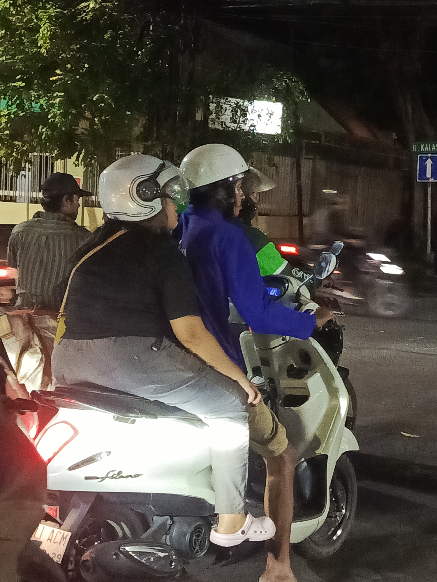 In Surabaya, Indonesia, at night, a group of individuals is depicted on a street. A person in the foreground, wearing a black t-shirt and grey pants, is seated on a white scooter. They are wearing a white helmet with black accents and a yellow strap across their chest. To their right, another person, wearing a blue jacket and a camouflage-colored helmet, is also seated on the scooter. Behind them, a third person, wearing a striped shirt and a dark baseball cap, is partially visible, appearing to be on a separate scooter. The scooter in the foreground has the license plate "L 1 ACM 29". The background shows trees, buildings, and street signs, including one that reads "Jl. Kalas." The scene is illuminated by artificial lighting, creating a night-time ambiance.