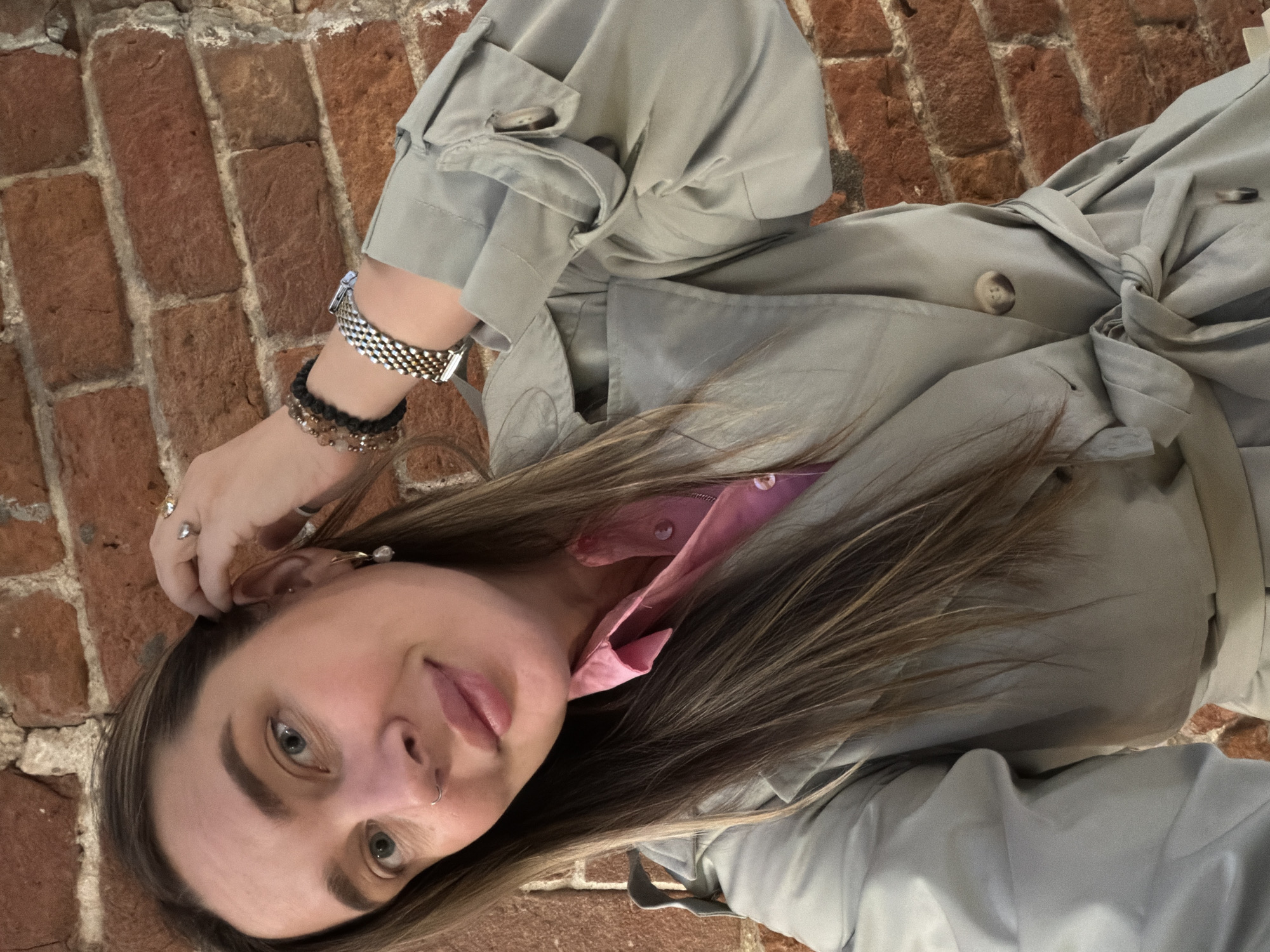 A young woman takes a selfie. She lies on a brick surface, possibly a sidewalk or building exterior. She is wearing a light green trench coat over a pink shirt. Her hair is brown with blonde highlights. She has a watch and bracelets on her left wrist, rings on her left hand, earrings, and a nose ring. The brickwork suggests an urban environment in Moscow, Russia.