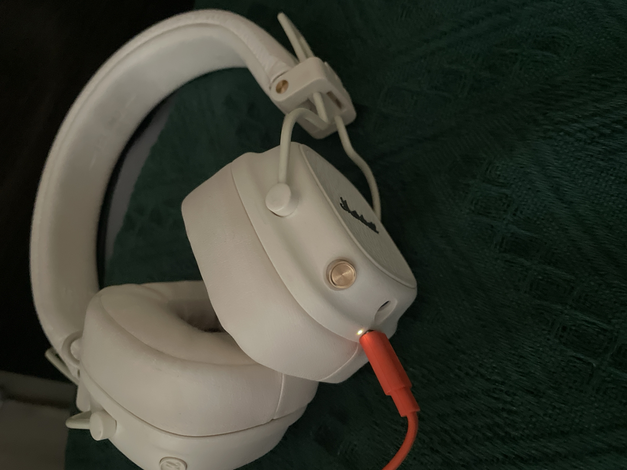 A pair of white Marshall headphones are resting on a dark green textured surface. The headphones are the primary object of focus. One ear cup is closer to the camera, showing a brass-colored button and an audio jack where an orange cable is plugged in. A small orange light is visible at the tip of the plugged-in cable, suggesting it's either charging or actively connected. The headband of the headphones is white and curved, with visible hinges and metal accents. The earcups appear to be made of soft material, likely for comfort. The overall setting is dimly lit, with the dark green surface providing a backdrop that contrasts with the white headphones. The image has a close-up perspective, highlighting the details of the headphones and the connection point. There is no indication of people or a specific time of day or weather conditions. The logo "Marshall" is visible in black lettering on the side of the ear cup.