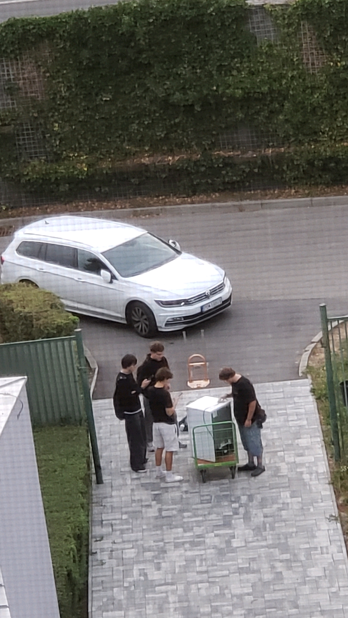 A group of four young males are gathered on a paved walkway next to a white Volkswagen station wagon parked on an asphalt road. The walkway is lined with a green metal fence on one side and a manicured hedge on the other. A small hand truck with a white appliance, possibly a refrigerator, is positioned between two of the individuals. One male, wearing a black t-shirt and denim shorts, is standing near the appliance, while another, dressed in a black hoodie and dark pants, is looking at it. Two other males, also in dark clothing, are standing slightly apart. A birdcage on a stand is visible in the background near the car. The scene appears to be in a residential or urban area, with a building visible in the foreground. The overall lighting suggests an overcast day. The location is in the city of Prešov, Slovakia.