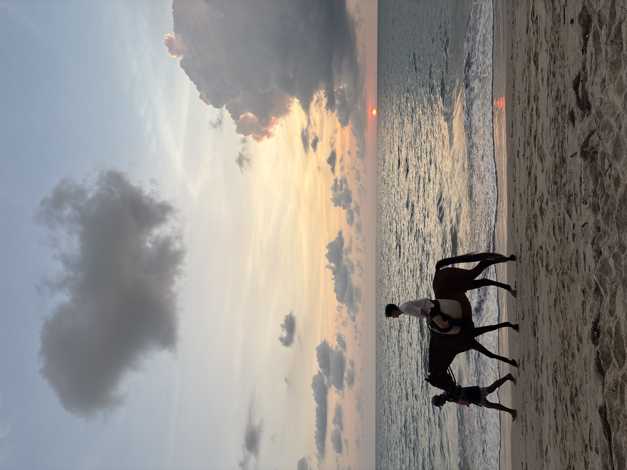 An outdoor image captures a serene sunset scene on a beach, likely in Choeng Thale, Thailand. The primary subjects are a person riding a dark brown horse, and another person walking alongside, leading the horse. The rider is clad in a light-colored (possibly white) long-sleeved shirt and dark trousers, seated in a saddle. The individual walking next to the horse wears dark shorts and a patterned shirt or vest.
The activity depicted is horseback riding along the coastline during the golden hour. The horse and people are positioned at the interface of the wet sand and the gently lapping ocean waves.
Notable details include the time of day: a vibrant sunset with the sun appearing as a clear orange orb low on the horizon, casting a warm glow across the sky and water. The sky transitions from pale blue overhead to warm orange, pink, and yellow closer to the horizon, dotted with scattered dark grey clouds, some with sun-kissed edges. The ocean reflects the sunset light, showing ripples and subtle wave patterns. The sandy beach displays natural textures and footprints from the people and horse. No visible text is present in the image.
