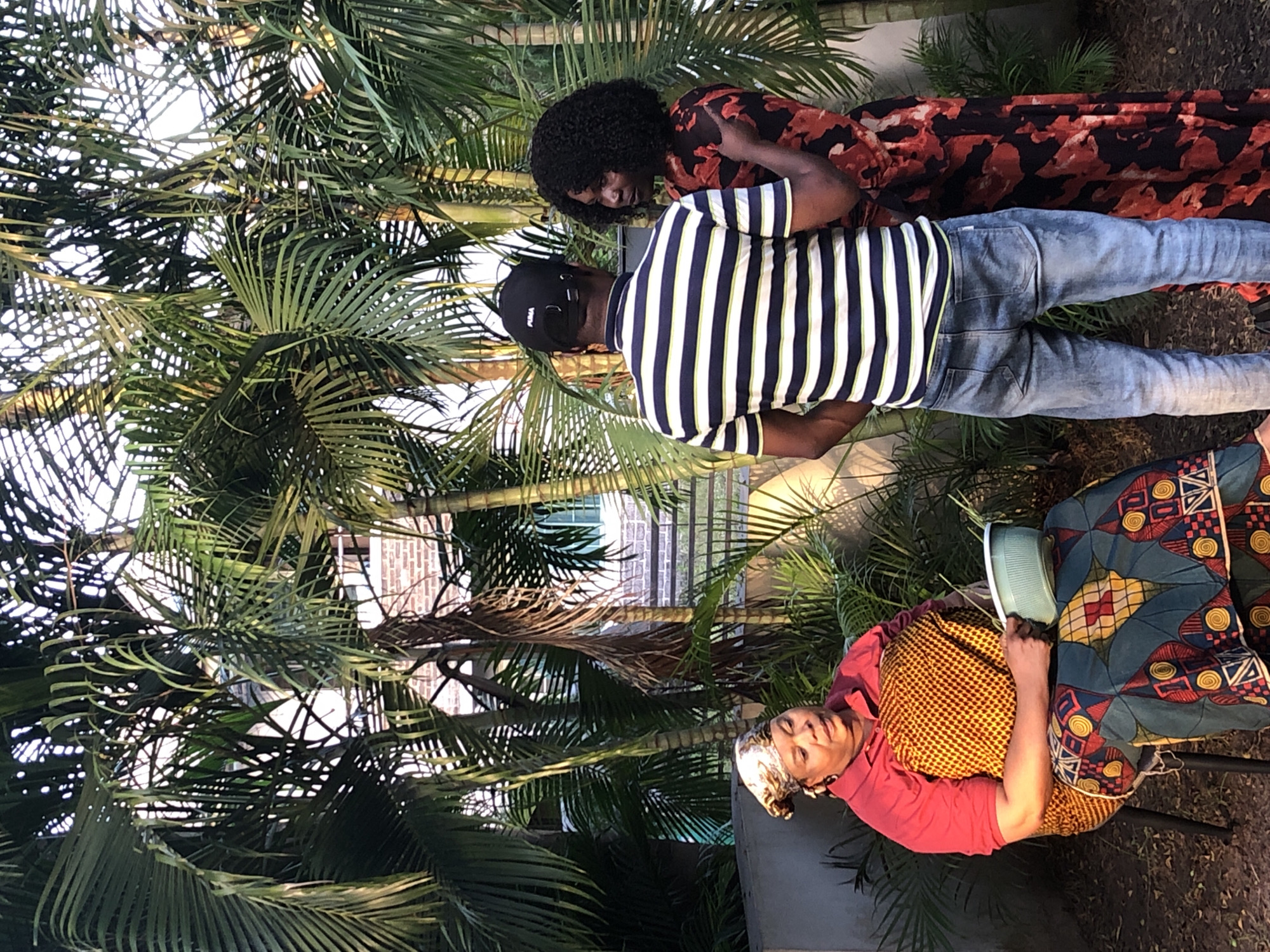 The image shows three people surrounded by palm trees. An older woman sits on a chair in the foreground, wearing a red shirt and a patterned yellow and blue skirt. She is holding a light green object. Behind her, a man in a striped shirt and jeans stands next to a woman in a floral dress. They appear to be embracing. A building is partially visible through the palm trees. The location is Unknown, Unknown.