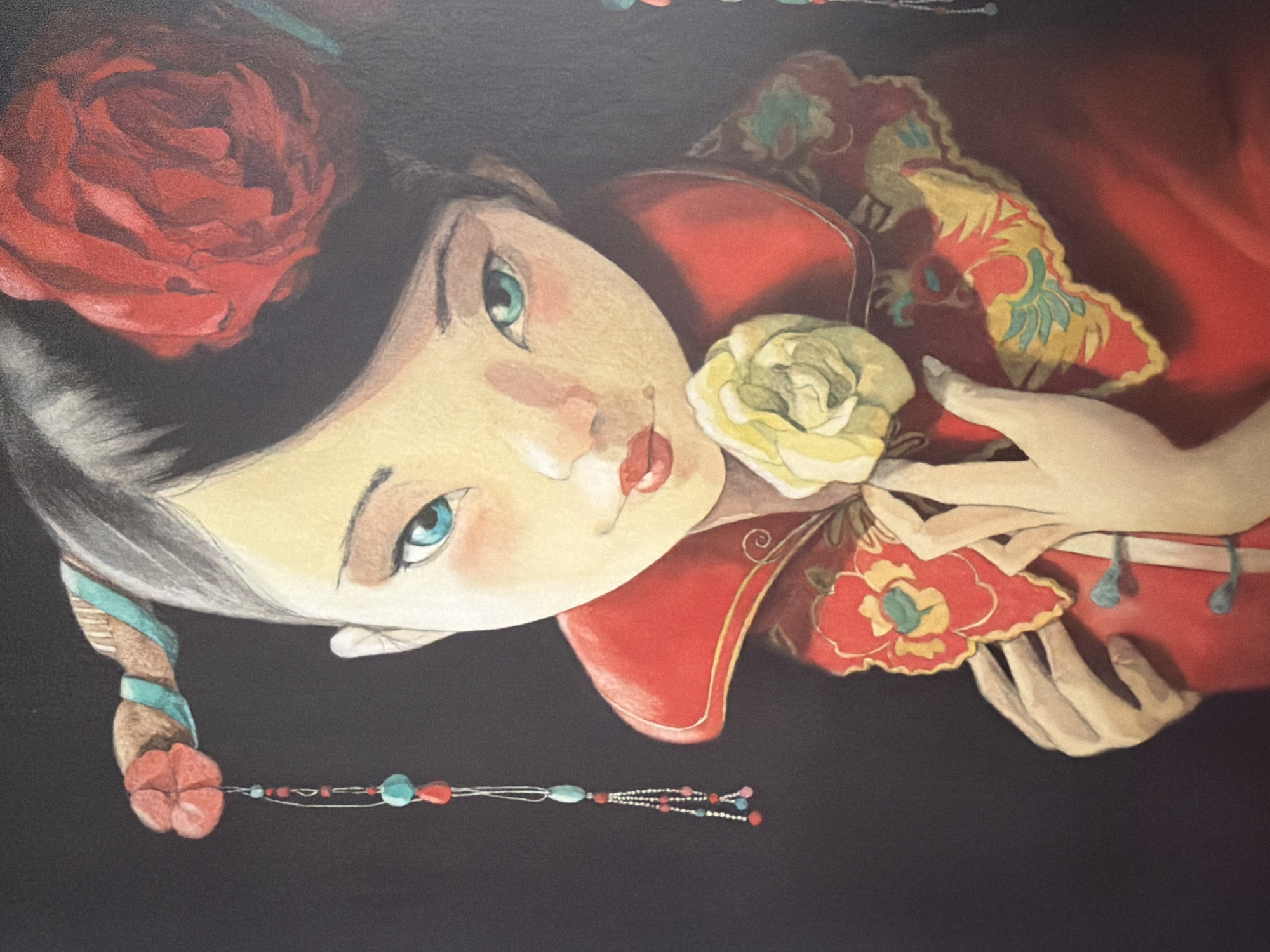 This is a painting of a young girl with striking blue eyes and pale skin, wearing traditional Chinese-inspired clothing. She has her hair styled in an updo with a large red flower and decorative hair accessories adorned with beads. Her garment is primarily red, with ornate gold and red floral embroidery on the collar and chest area. She is holding a pale yellow flower to her chest with her hands. The background is a solid dark color, making the subject stand out. There is no information provided about the painting's location in Pazardzhik, Bulgaria.