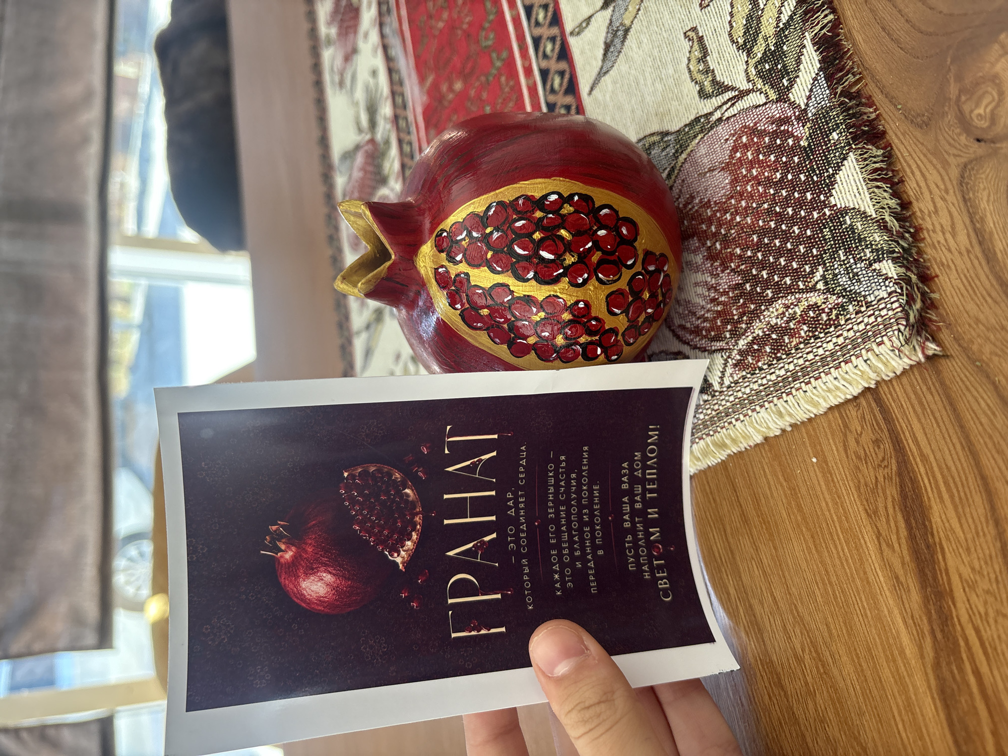 A hand holds a postcard-like item featuring an image of a pomegranate with text in Russian. The text translates to "POMEGRANATE - IT IS A GIFT THAT UNITES HEARTS. EACH SEED IS A PROMISE OF HAPPINESS AND WELL-BEING, PASSED DOWN FROM GENERATION TO GENERATION. MAY YOUR VASE FILL YOUR HOME WITH LIGHT AND WARMTH!" Next to the postcard, on a patterned tablecloth, sits a decorative pomegranate figurine. The scene appears to be indoors, with natural light filtering in from a window in the background, where a blurred view of trees and possibly a building can be seen. The table is made of wood. The provided information does not include location details for Georgiyevsk, Russia.