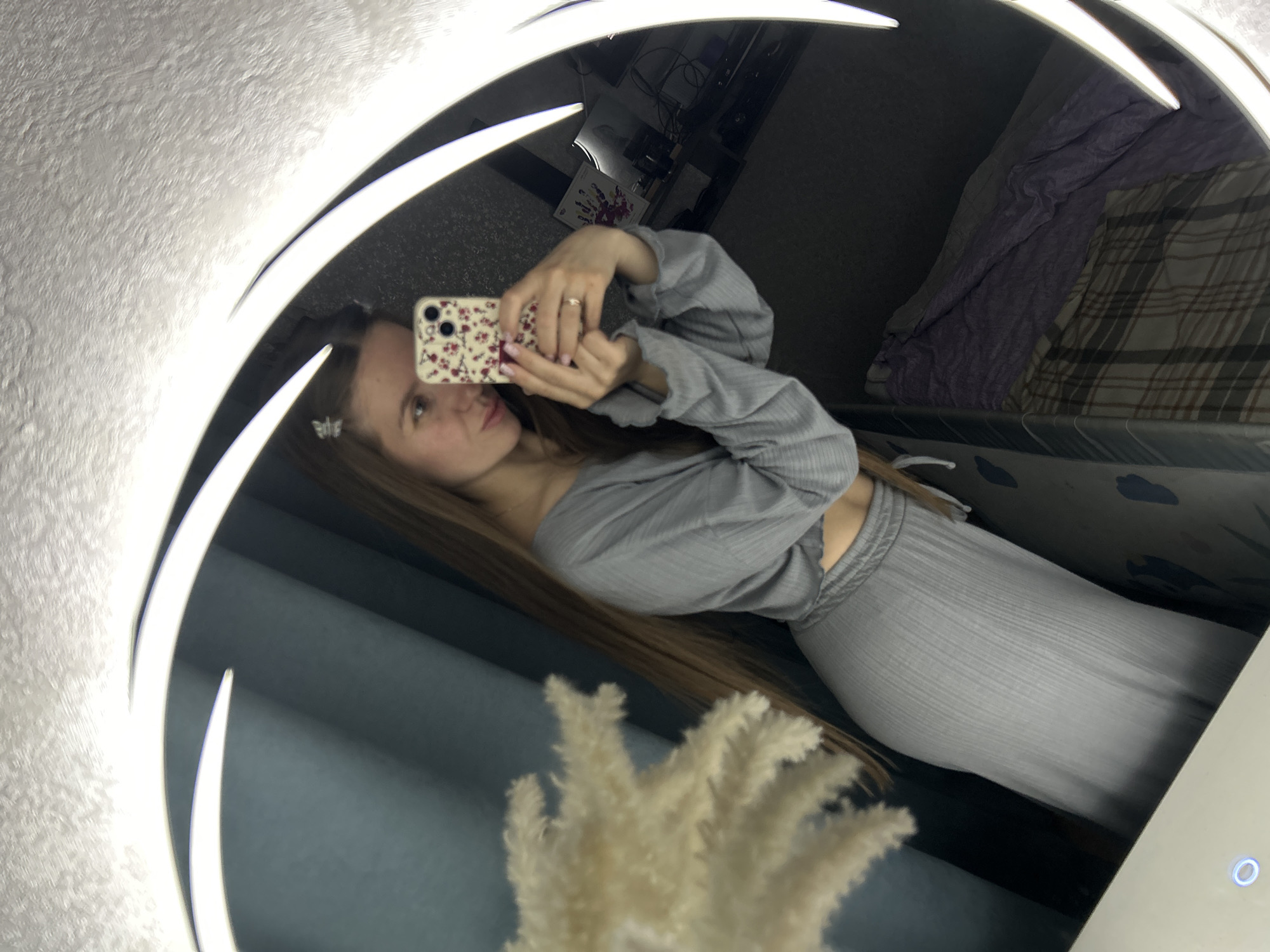 A young woman is captured in a mirror selfie, holding a smartphone with a floral-patterned case featuring red and white flowers on a light background. She is wearing a light grey, ribbed two-piece outfit composed of a long-sleeved crop top and matching pants. A ring is visible on her right ring finger, and a small, butterfly-shaped hair clip adorns her hair on her right side.
The image is taken through a large, possibly circular or oval, mirror that has integrated bright white LED lighting around its edges. In the immediate foreground, at the bottom center, a fluffy, light-colored decorative plant or arrangement, possibly pampas grass, is partially visible and blurred.
The reflection in the mirror reveals an indoor setting. To the left and top, a textured white or light-colored wall can be seen. In the upper right corner of the reflection, there is a bed or sofa covered with a purple textured blanket and a plaid blanket. Further in the background, behind the woman's left shoulder, appears to be dark furniture or shelves with various objects. A blue circular indicator light is visible on a white surface in the bottom right of the mirror's reflection. The strong illumination from the mirror suggests the photo was taken indoors, possibly in a bedroom or dressing area. No discernable text is visible.