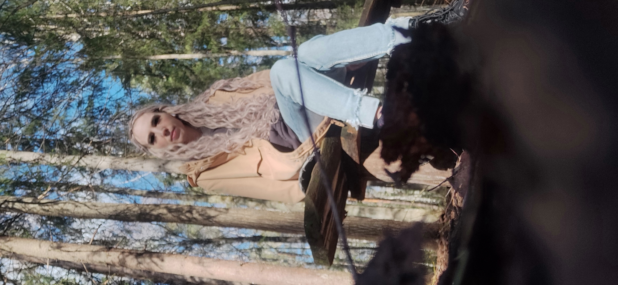 The image shows a person with long, curly blonde hair, wearing a tan jacket and light blue jeans, sitting on a wooden structure in a wooded area. Trees surround the person, with glimpses of a blue sky visible through the branches. The location and city are not evident from the image. A dark, blurred object is present in the foreground, partially obscuring the view.