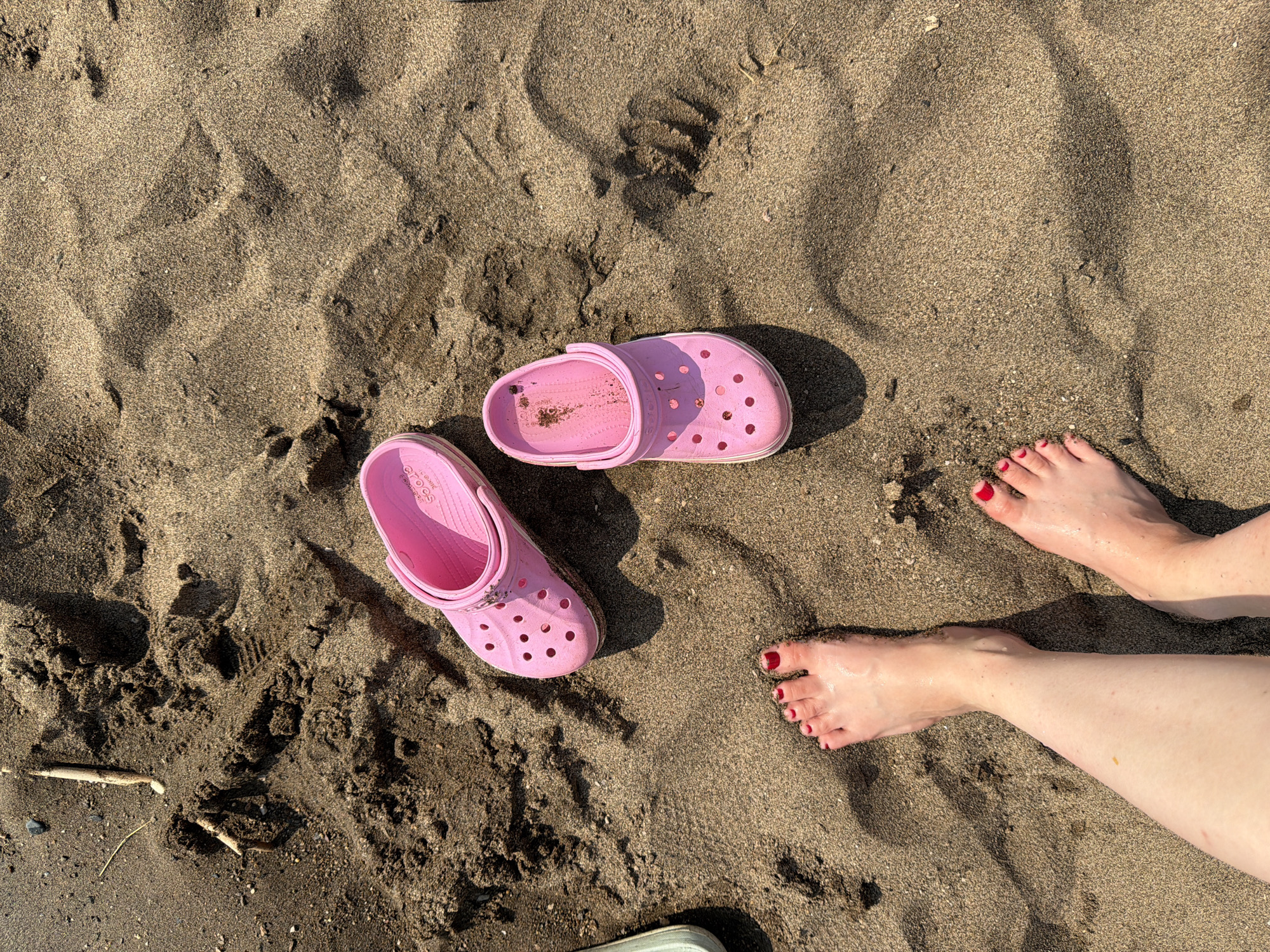 The image displays a top-down view of sand on a beach, with two pink Crocs shoes and two feet. One foot, with red painted toenails, is visible on the lower left, with its heel pressed into the sand. The other foot, also with red painted toenails, is positioned further down and to the right. One pink Croc is on the left side of the frame, and another is on the right. The sand is dark brown and appears wet in places, with small debris and indentations indicating footprints. The scene is likely an outdoor location, possibly a beach or a riverbank in Krasnyi Bor, Russia, given the context provided. The lighting suggests it is daytime, casting shadows on the sand.
