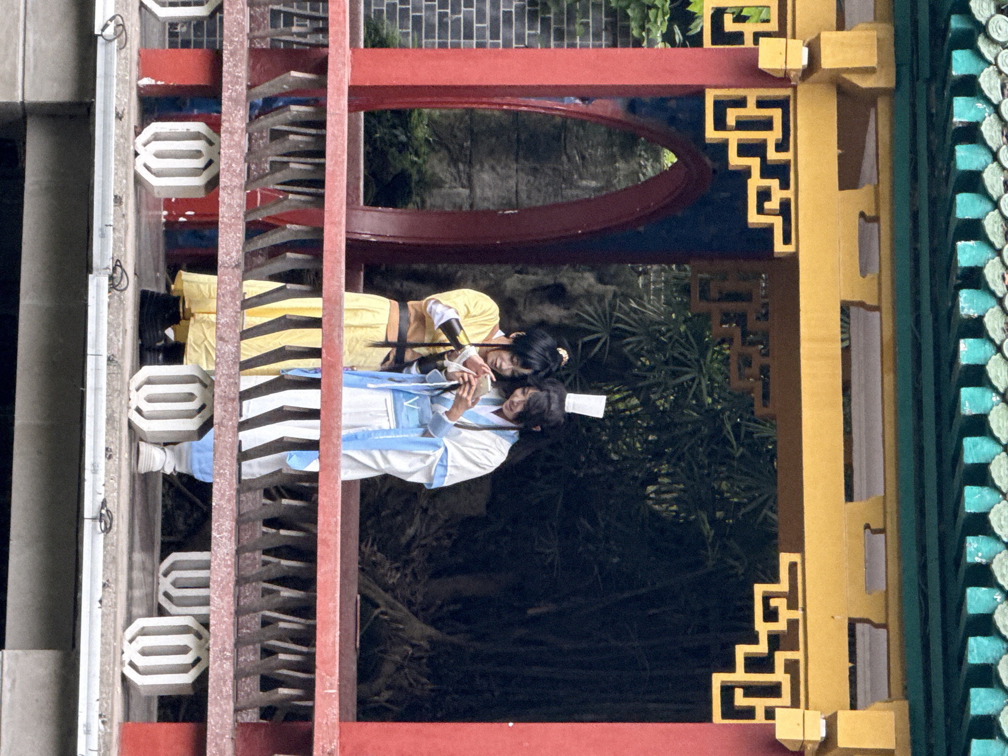 Two people, one in yellow and one in white and blue traditional Chinese clothing, are positioned in front of a red and yellow ornate archway structure. The red structure has a decorative oval opening at the top, while the yellow structure features an intricate geometric design. Greenery can be seen through the archway. In the background, there is a brick wall and additional foliage. The people appear to be looking at or interacting with a mobile phone. The location is possibly in Yuzhong District, China, based on the traditional Chinese architectural style.
