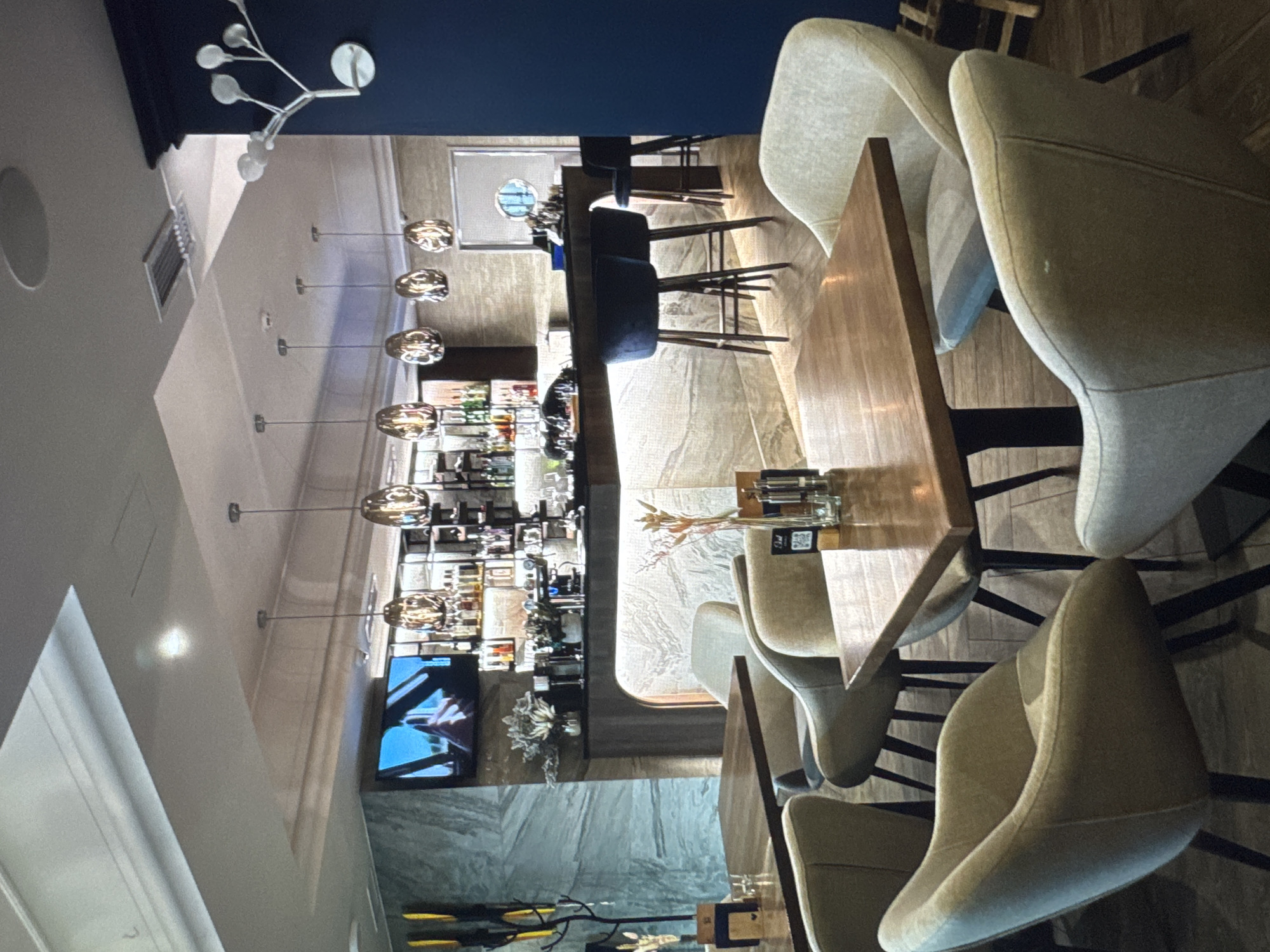 The image depicts the interior of a modern, well-lit restaurant or bar. The scene is dominated by seating areas with light-colored upholstered chairs and dark wooden tables. In the background, a long bar counter is visible, stocked with bottles and illuminated from below. Several contemporary pendant lights hang above the bar. A television screen displays an image of a person driving. The walls are decorated with a patterned marble-like finish and a dark blue accent wall. On the far right, a decorative wall sconce resembling a tree branch with lights is mounted. The flooring appears to be dark wood. The overall impression is one of a sophisticated and comfortable dining or drinking establishment. The location, Новинки, Russia, is a small village in the Nizhny Novgorod Oblast, suggesting this establishment is a local amenity rather than a large city venue.