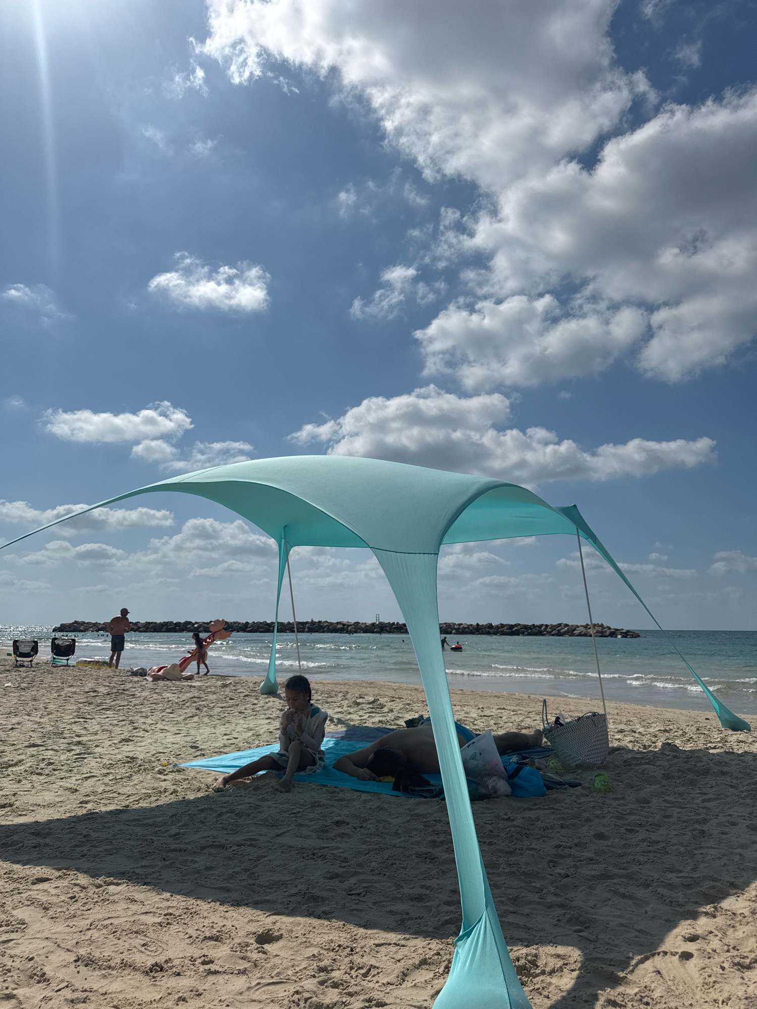 The image depicts a sunny day at a beach in Netanya, Israel, likely along the Mediterranean coast. A light blue beach tent is set up in the foreground, providing shade. Underneath the tent, a young child sits on a blue mat, holding something to their mouth, while an adult lies down next to them. Nearby, a patterned beach bag and some other objects are visible on the sand. In the middle ground, several people are scattered on the beach. A man stands near the water's edge, facing away from the camera, while other individuals are lying down or walking on the sand. In the background, the sea is calm with gentle waves, and a breakwater made of large rocks extends into the water. The sky is a bright blue with scattered white clouds, and the sun is shining brightly, casting shadows on the sand. The overall scene is one of leisure and relaxation at the beach.