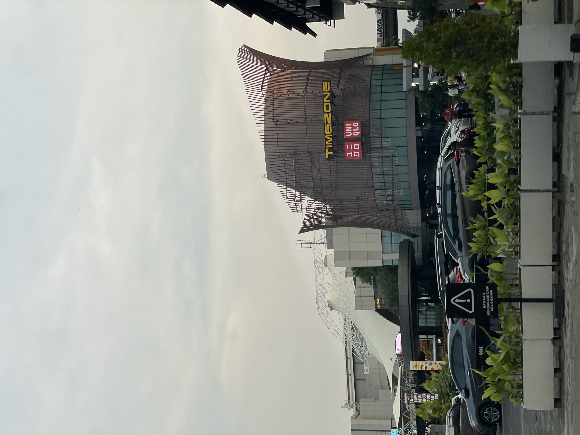 The image depicts an urban scene in North Jakarta, Indonesia, showcasing a Timezone arcade and Uniqlo store within a multi-story complex. The building's facade features a distinctive metal framework. Several parked cars are visible in the foreground. A warning sign with a red exclamation point is also present, cautioning about vehicles approaching from the right. The sky is overcast.