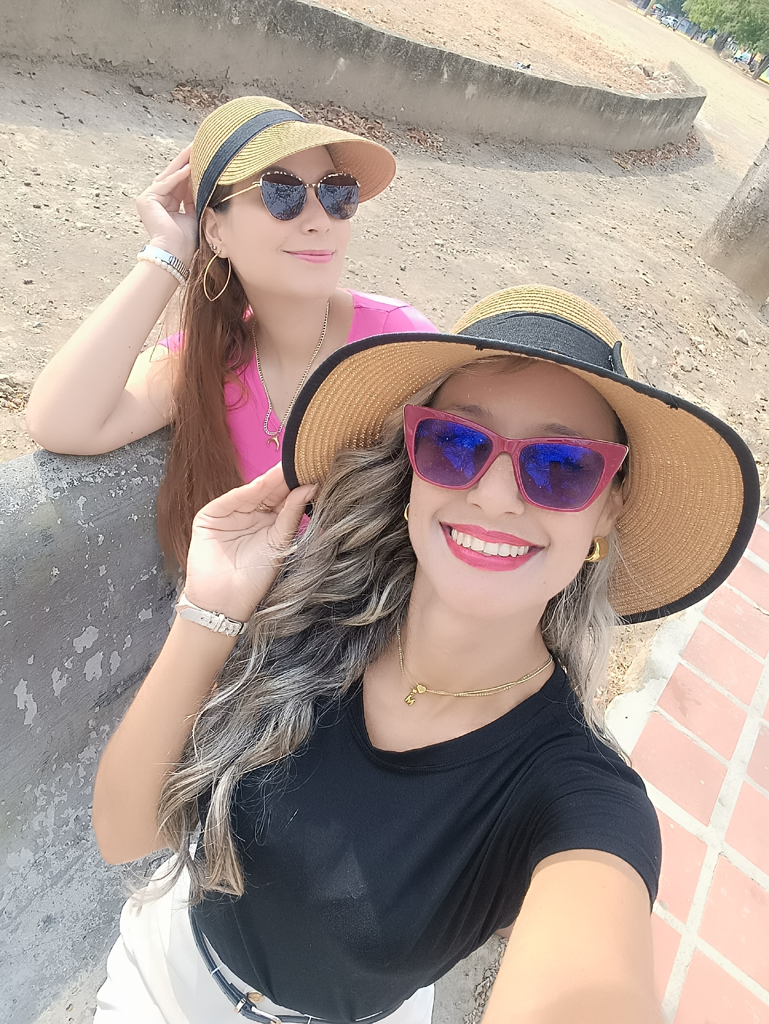 Two adult females are depicted in an outdoor setting in La Victoria, Venezuela, during daylight hours. The subject in the foreground is taking a selfie, facing the camera with a smile. She wears a wide-brimmed straw hat with a black band, red-framed sunglasses with reflective blue lenses, gold hoop earrings, a gold choker necklace with a small pendant, a black short-sleeved top, and white lower garment. Her hair is wavy with light-colored highlights.
Behind and to the left, a second female subject is partially visible, smiling and looking to her right, away from the camera. She wears a straw hat with a black band, dark cat-eye sunglasses, gold elongated hoop earrings, a gold chain necklace with a crescent moon pendant, and a pink short-sleeved top. Her hair is long and brown. Her left forearm rests on a weathered, low concrete structure, and her right hand is near her hat.
The immediate ground level to the right of the foreground subject is reddish-brown brick pavement. The area surrounding the subjects and extending into the background consists of dry, light-colored earth or sand with scattered dry leaves. A low, curved concrete wall or bench, showing signs of wear and peeling surface, is prominent in the middle ground. The distant background features blurred green foliage and indistinct structures, including what appear to be parked vehicles, suggesting an open public or semi-urban area.