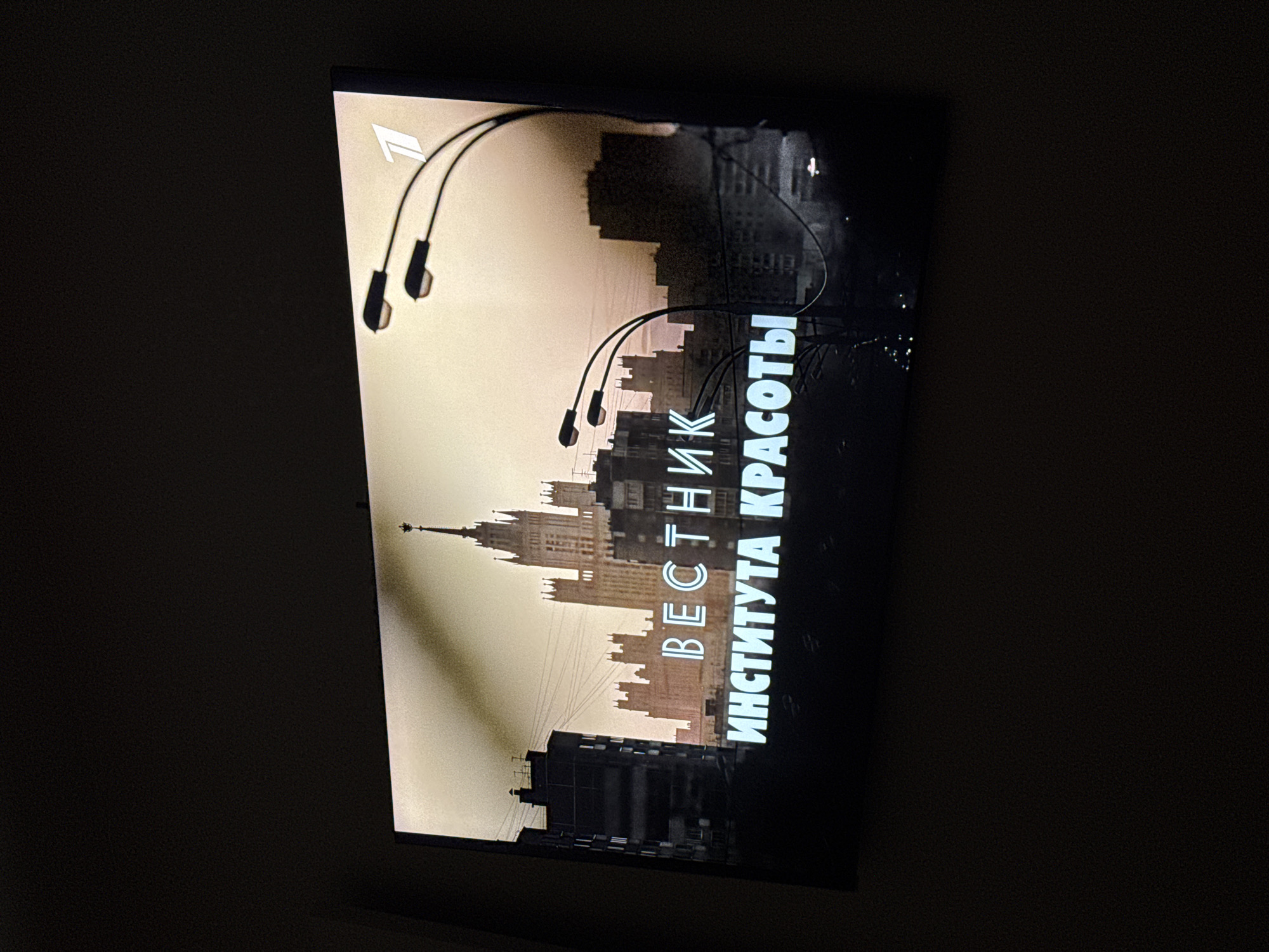 A television screen displays a scene from "Vestnik Krasoty Instituta" set in the city of Unknown, Unknown. The image is in sepia tones. In the foreground, the title is written in Russian. Above the text, the skyline of a city is visible, dominated by a tall, spire-topped building reminiscent of Soviet architecture. In front of this building and dispersed, are a number of street lamps. The illumination on the screen is brighter on the left side, fading into darkness on the right. The surrounding environment is dark.